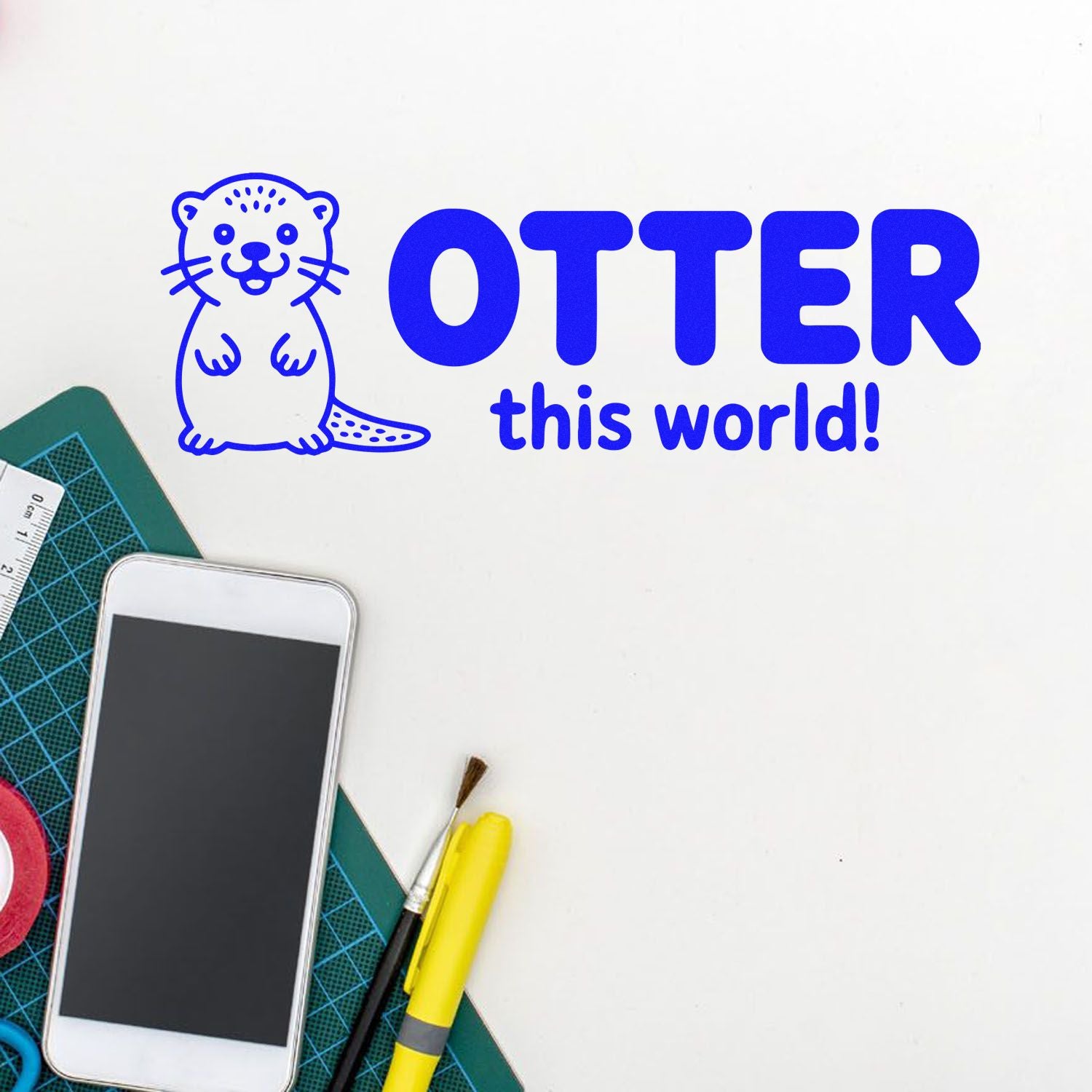 Blue otter stamp and seal design with OTTER this world! on white, beside craft tools and cutting mat.