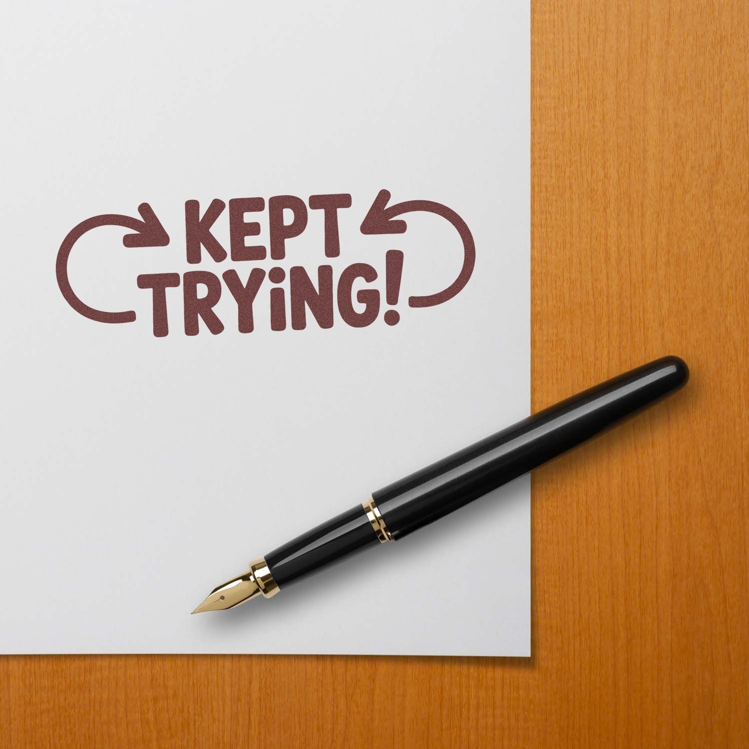 Maroon Kept Trying! rubber stamp with arrows, black fountain pen, on white paper over a wood desk.