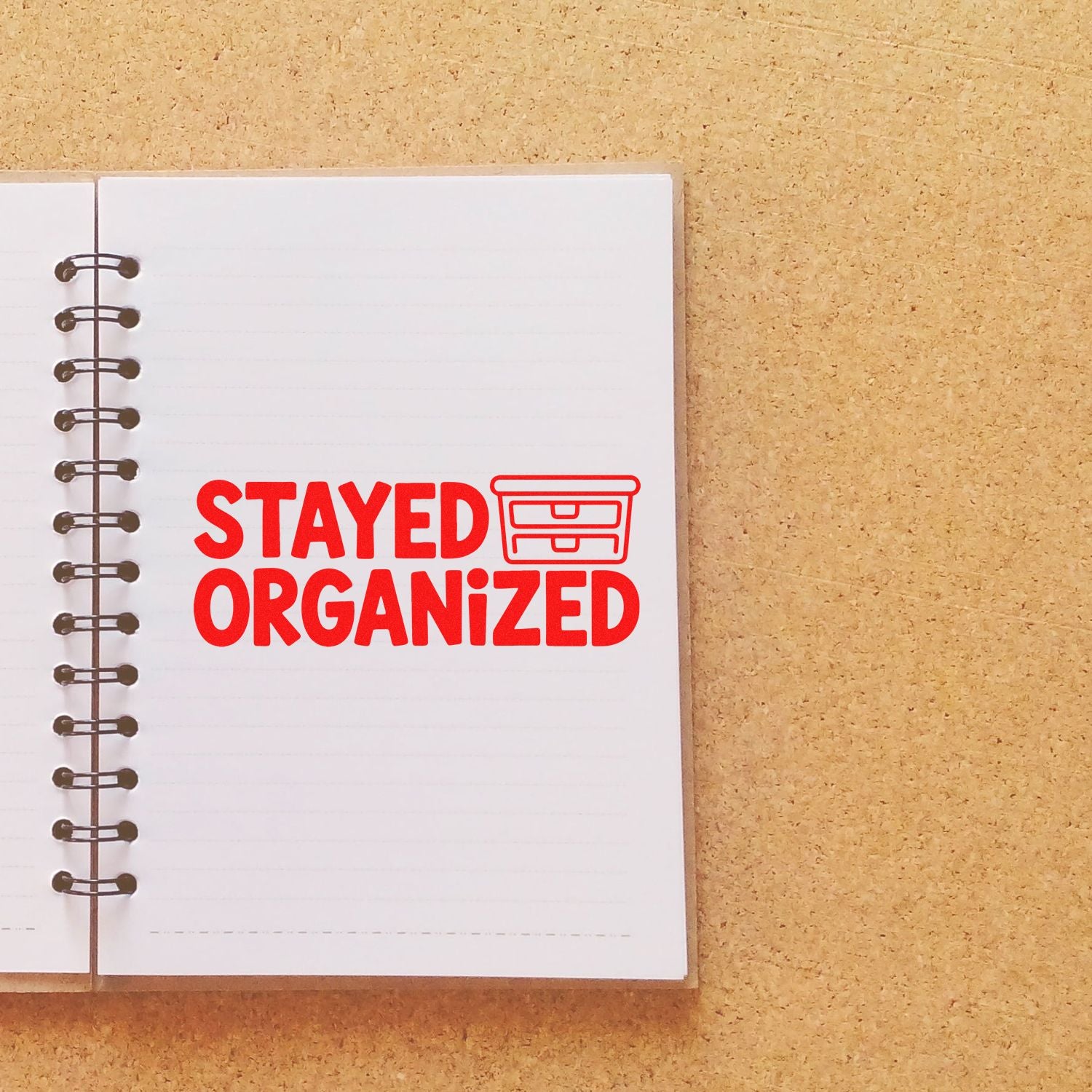 Red STAYED ORGANIZED stamp and seal with drawer icon on a white spiral notebook over tan cork background.