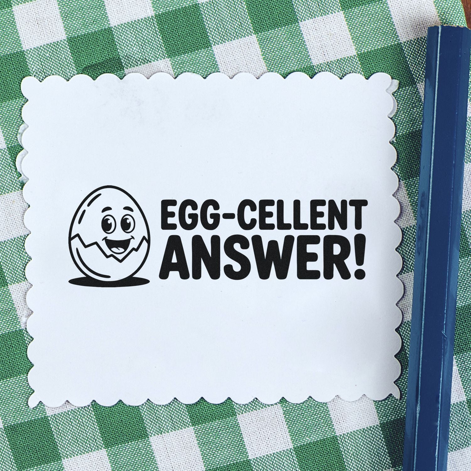 White scalloped stamp/seal card with black egg icon and EGG-CELLENT ANSWER on green checkered cloth, beside blue pen.