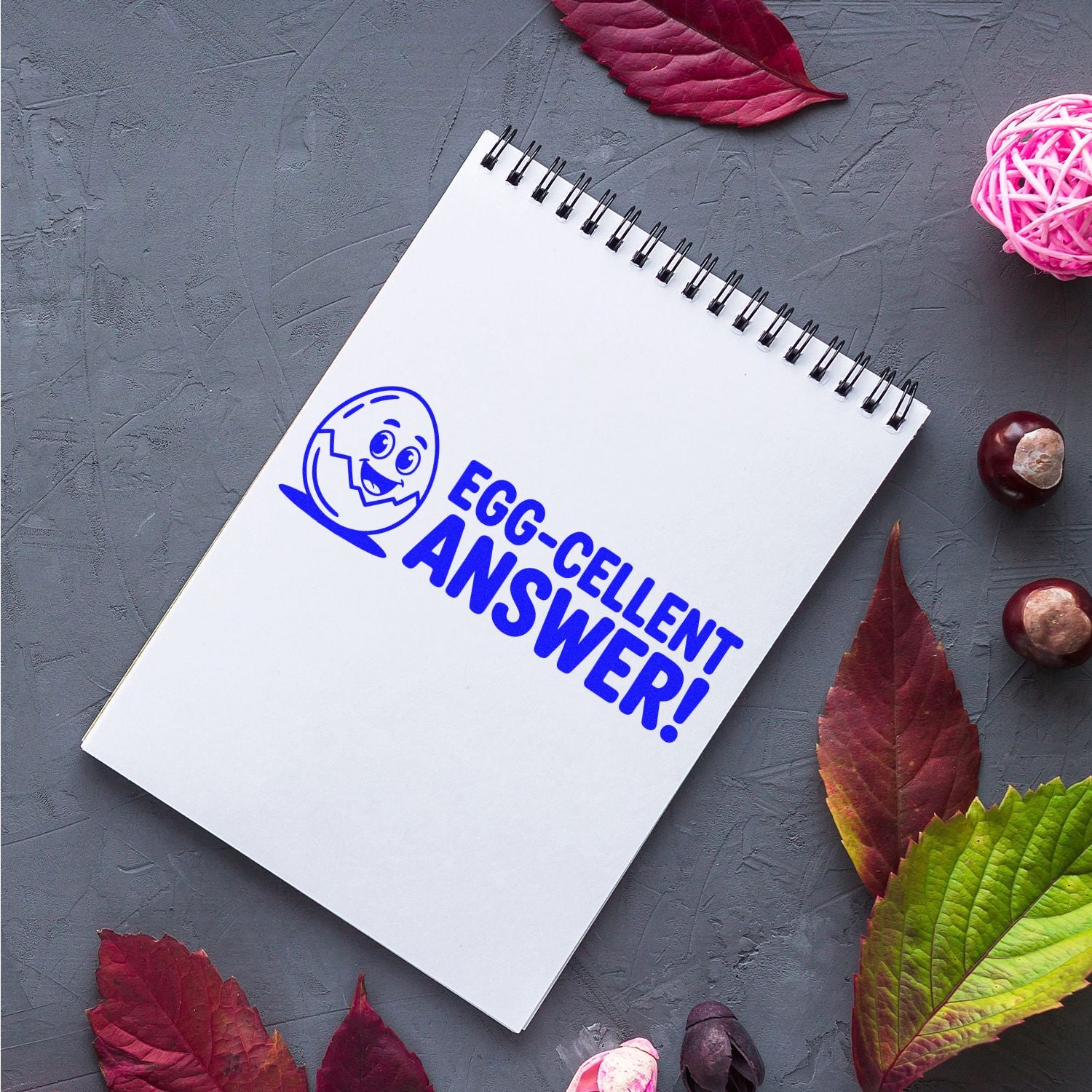Blue egg-themed stamp and seal reading EGG-CELLENT ANSWER! on white spiral notebook, with autumn leaves.