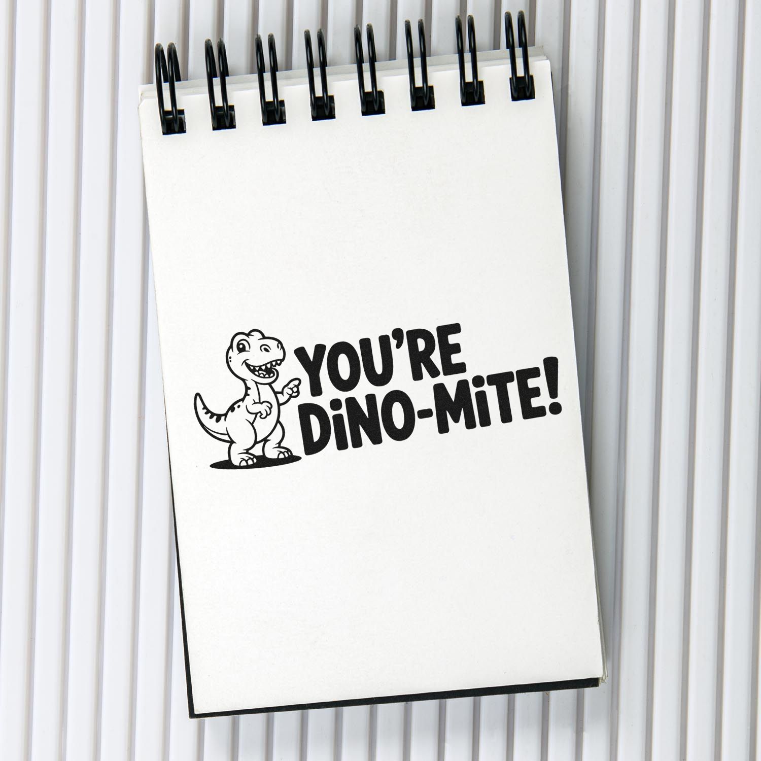 White spiral notebook with a black dino stamp reading YOURE DINO-MITE! on a light gray background.