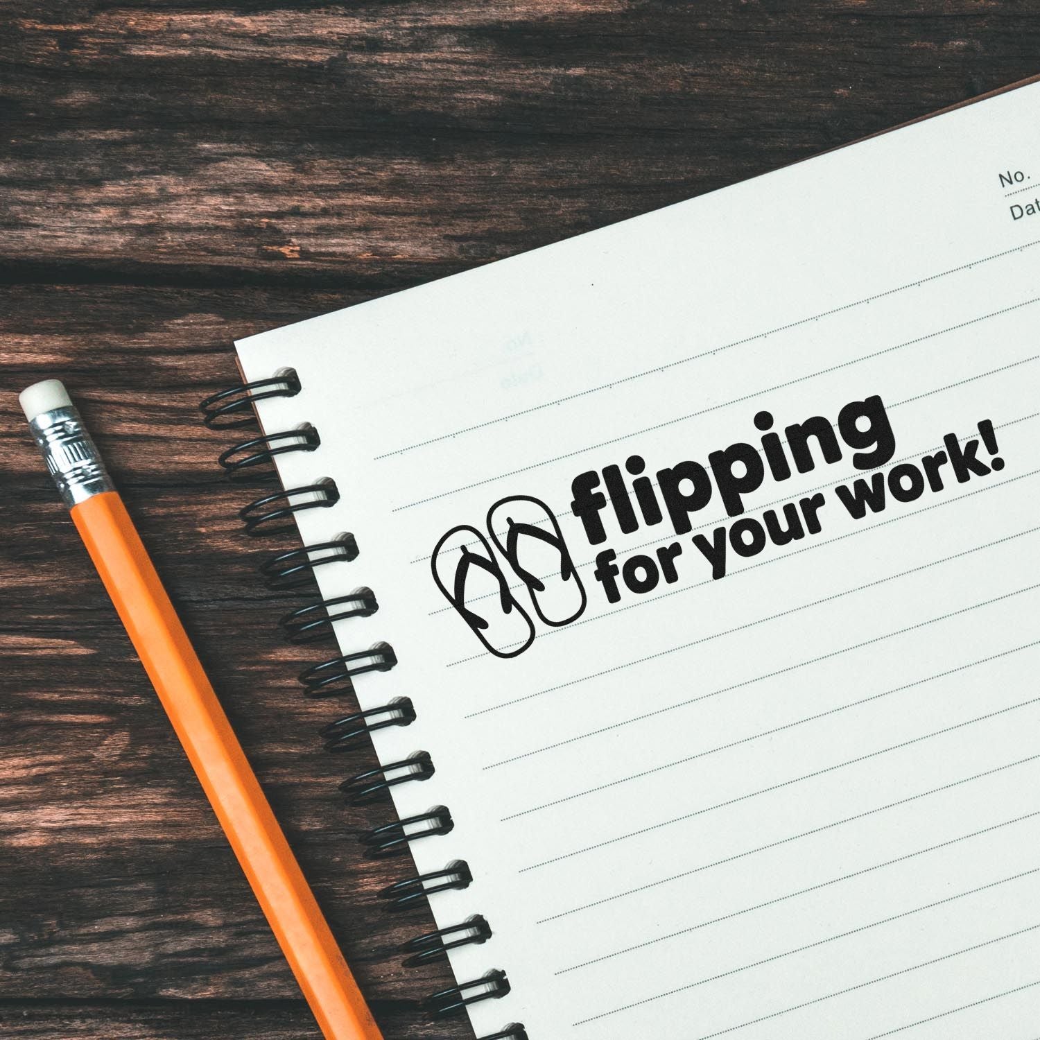 Black custom stamp impression reading flipping for your work! on white spiral notebook, with orange pencil on dark wood.