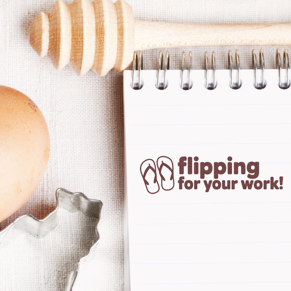 Brown flip-flop stamp seal on white spiral notebook, with wooden honey dipper, egg, and metal cutter on beige fabric.