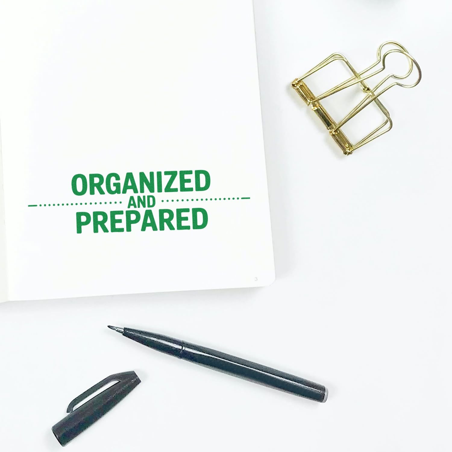 White notebook with green Organized and Prepared stamp, gold binder clip, and black pen on a clean white background.
