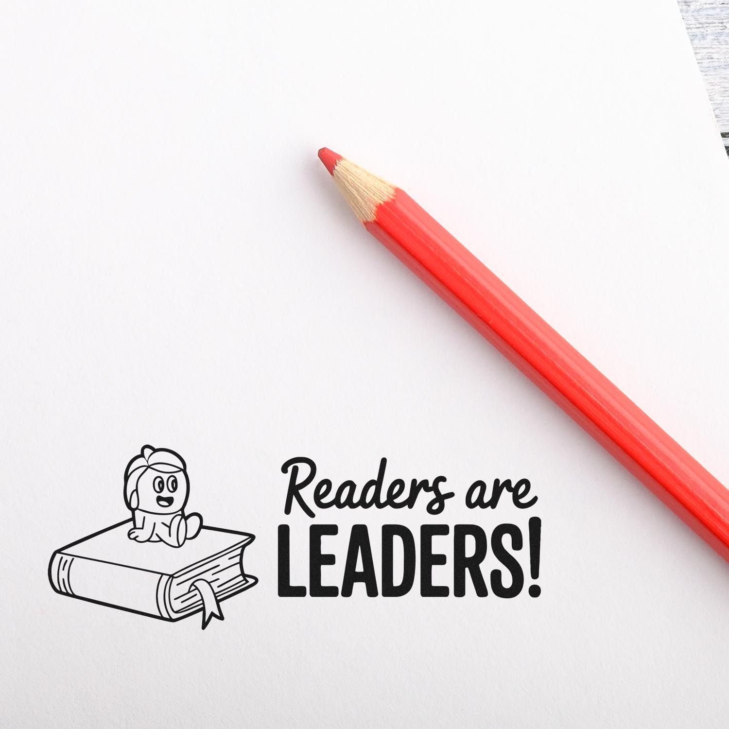 Red pencil on white paper beside a black reading-themed stamp seal with cartoon child on a book and Readers are LEADERS!