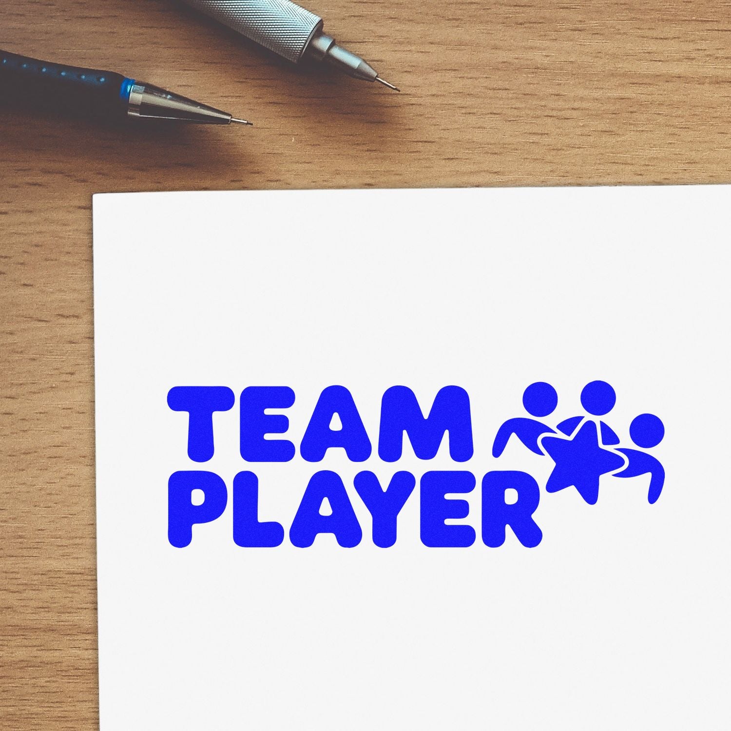 Blue TEAM PLAYER stamp and seal on white card, with people-and-star icon, beside pens on a wooden desk.