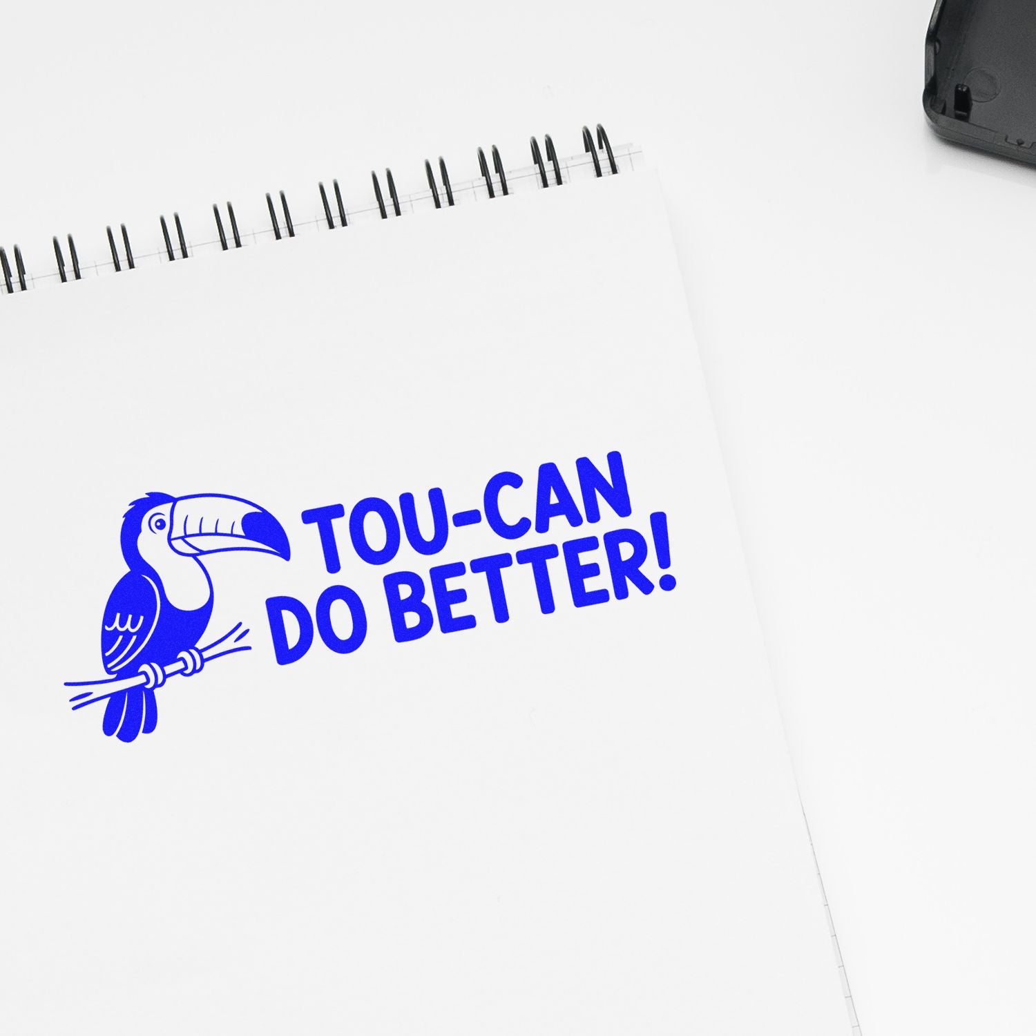 Blue toucan stamp on white spiral notebook with bold TOU-CAN DO BETTER! text; black stamp case visible in corner.