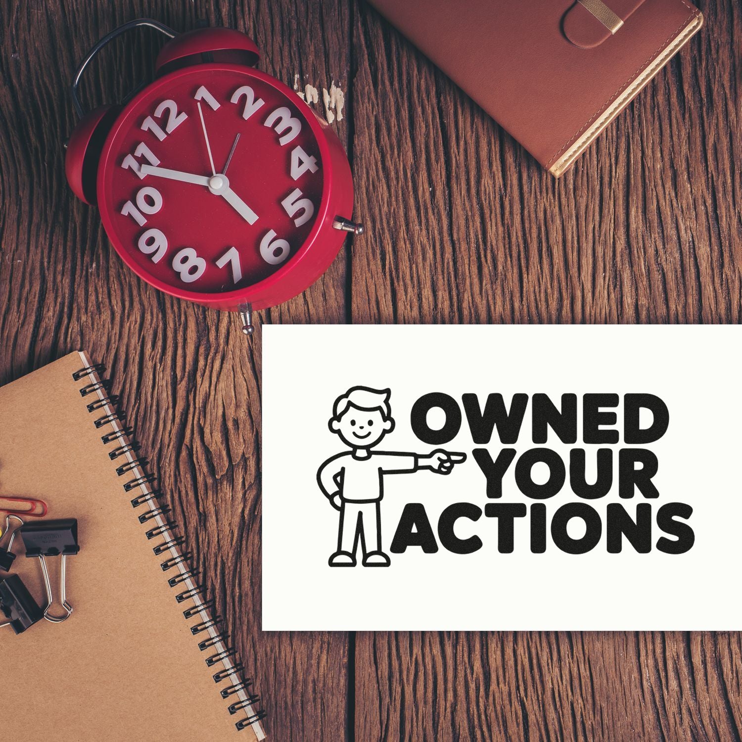Black OWNED YOUR ACTIONS stamp and seal on white card beside a red alarm clock on a wood desk.