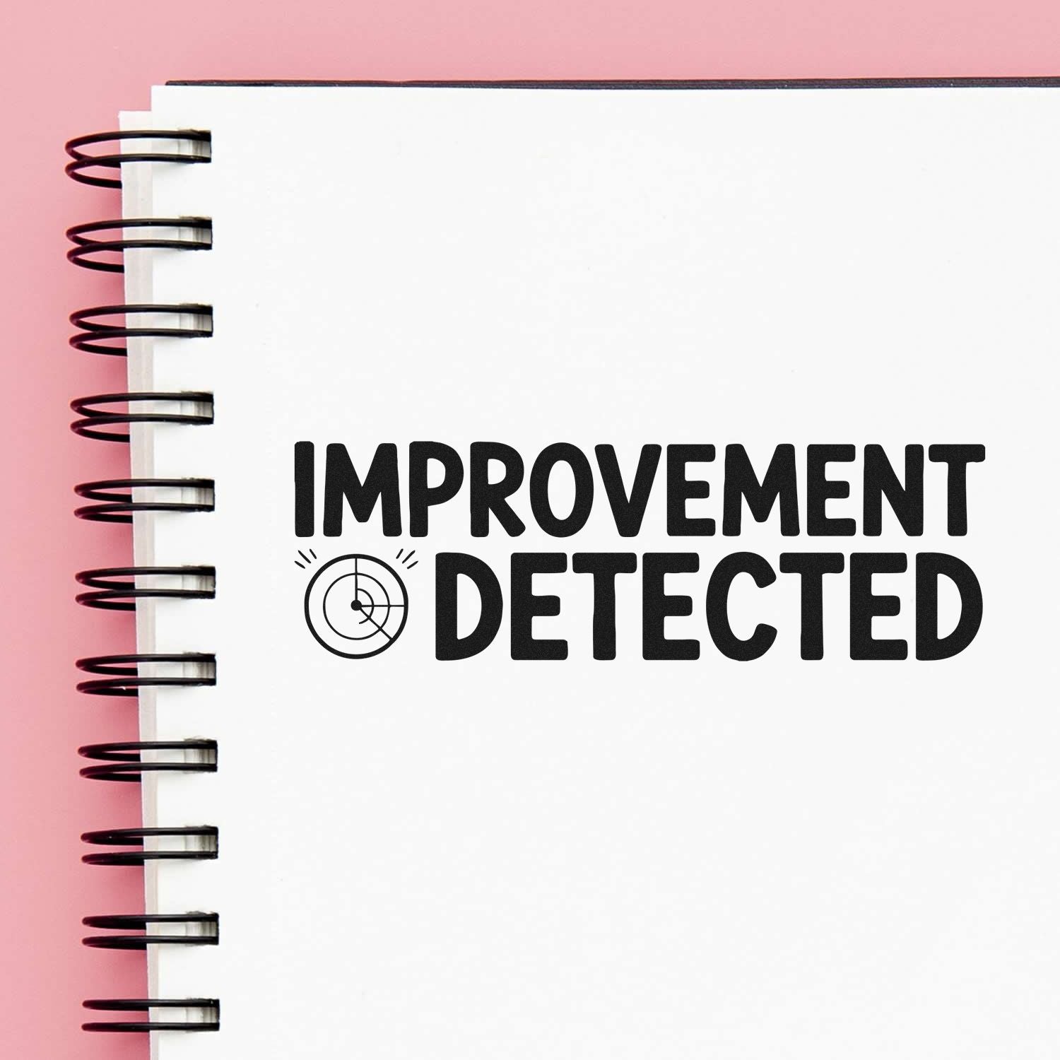 White spiral notebook with black stamp-style seal reading IMPROVEMENT DETECTED and target icon on pink background.