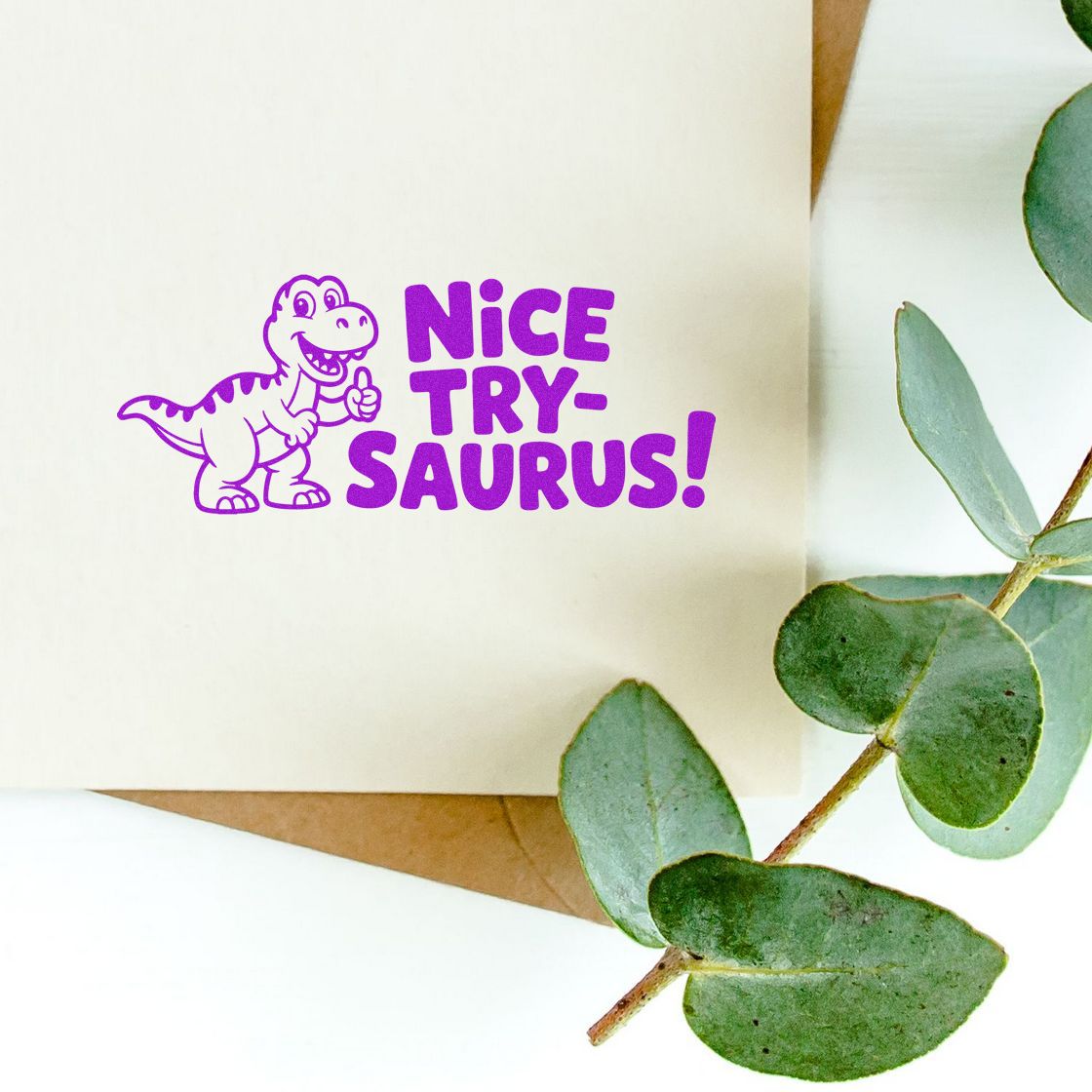 Purple dinosaur stamp reading Nice Try-saurus! on cream paper, with green eucalyptus leaves nearby.