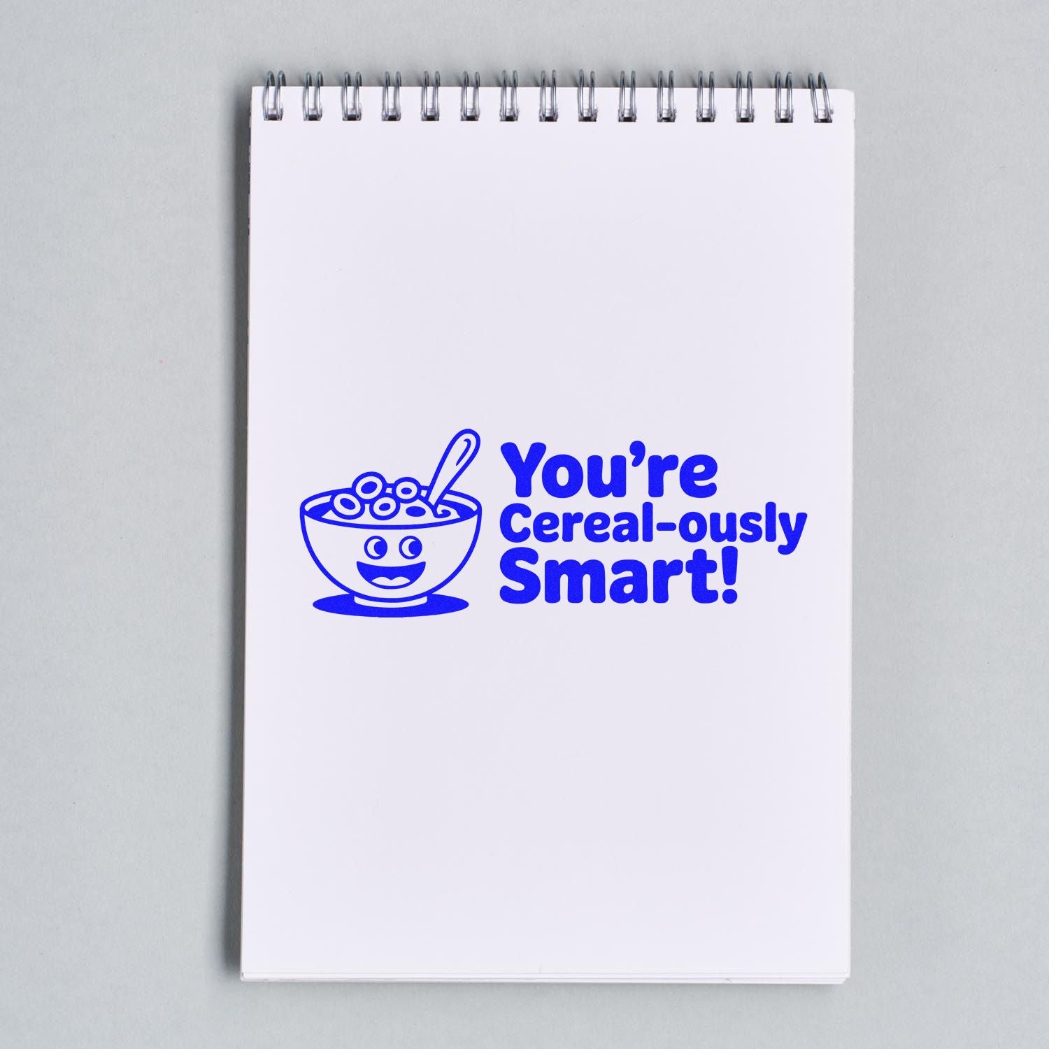 Blue self-inking stamp on a white spiral notepad, showing a cereal bowl and text: Youre Cereal-ously Smart!