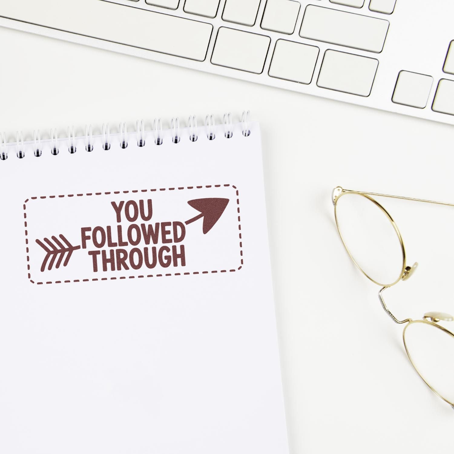 Brown You Followed Through rubber stamp seal on white notepad beside a white keyboard and gold glasses.