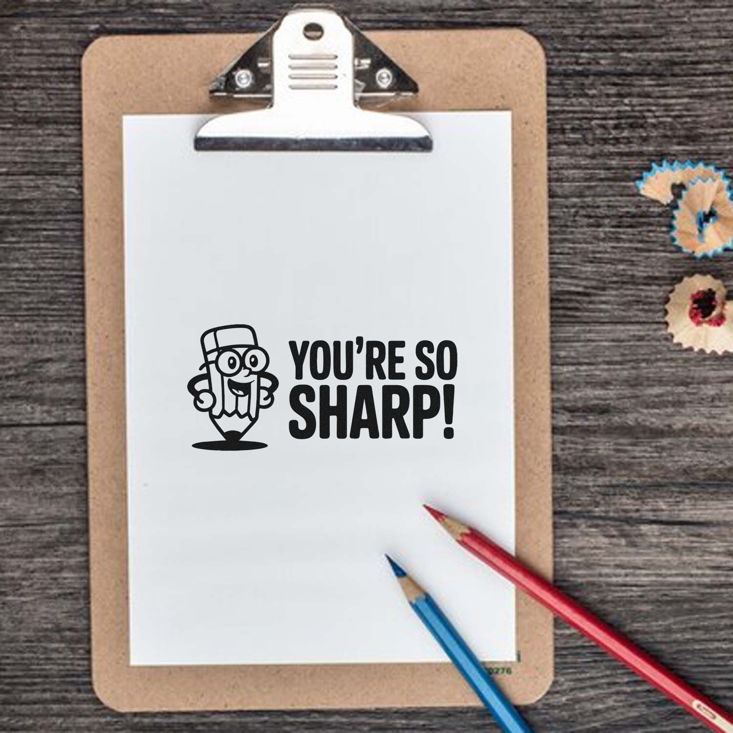 Black Youre so sharp! pencil stamp on white clipboard, with red and blue pencils and colorful seal stickers on wood.