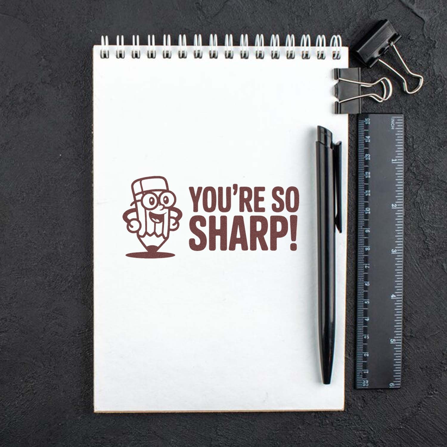 Brown self-inking stamp reading YOURE SO SHARP! with cartoon pencil on a white notebook, beside a black pen, ruler, and binder clips.