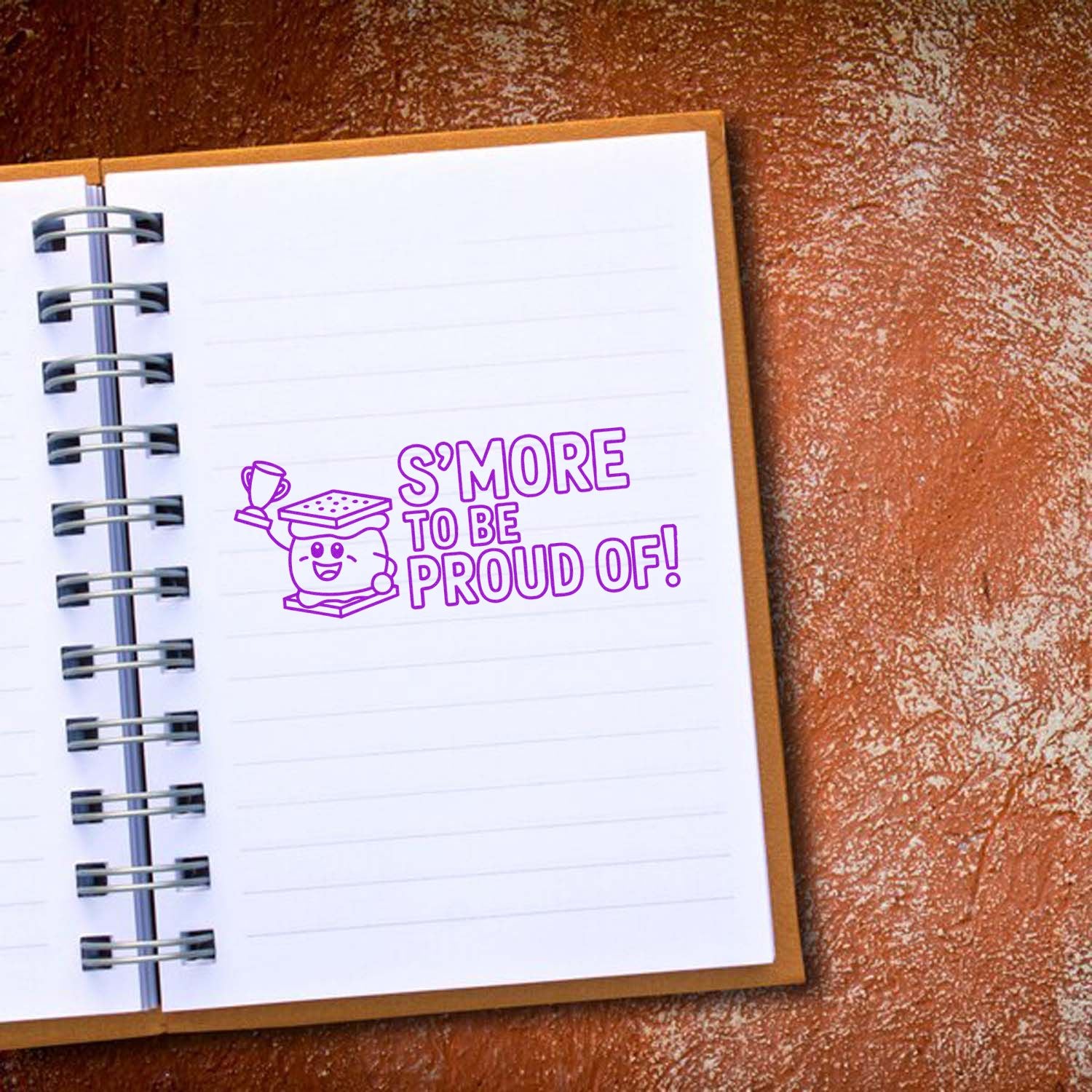 Purple stamp seal with graduate mascot and SMORE TO BE PROUD OF! on a lined notebook.