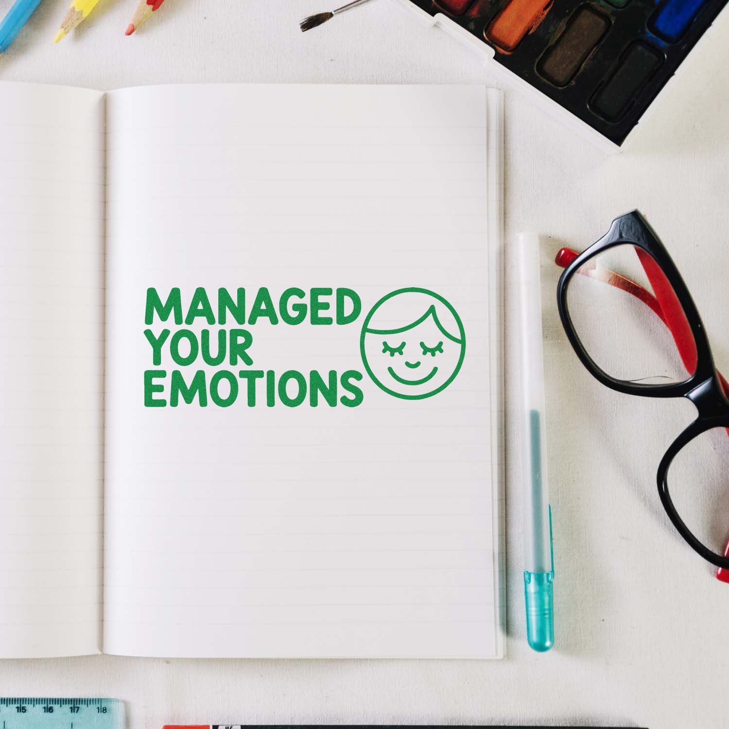 Green self-inking stamp seal reading "Managed Your Emotions" on an open notebook, with smiling face icon, glasses, and art supplies.