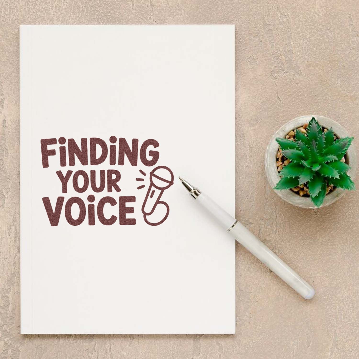 White notebook stamped with maroon Finding Your Voice and microphone seal, with a white pen and green succulent.