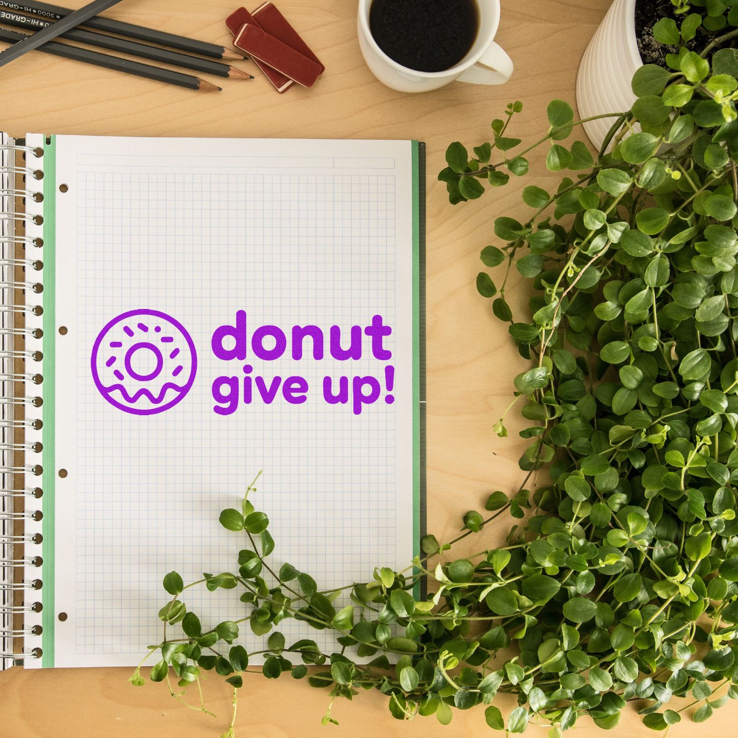 Purple donut give up! stamp and seal design on graph paper notebook, with pencils, coffee, and green plant.