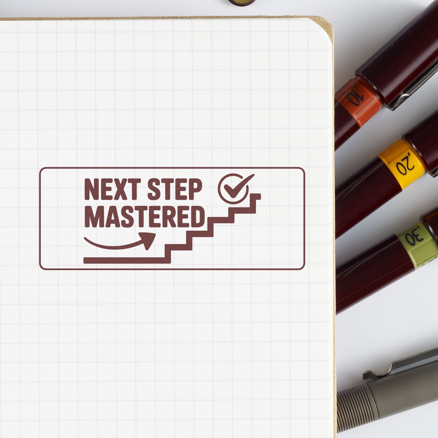 Brown stamp seal reading Next Step Mastered on grid paper, beside burgundy markers with orange, yellow, and green seals.