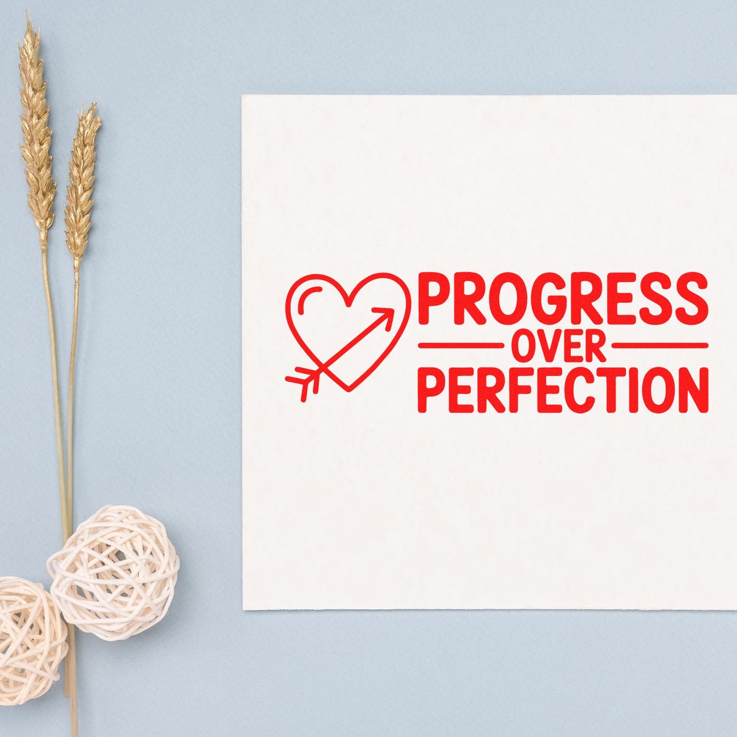 Red progress over perfection stamp seal with heart arrow on white paper, set against a light blue background with gold wheat stems.