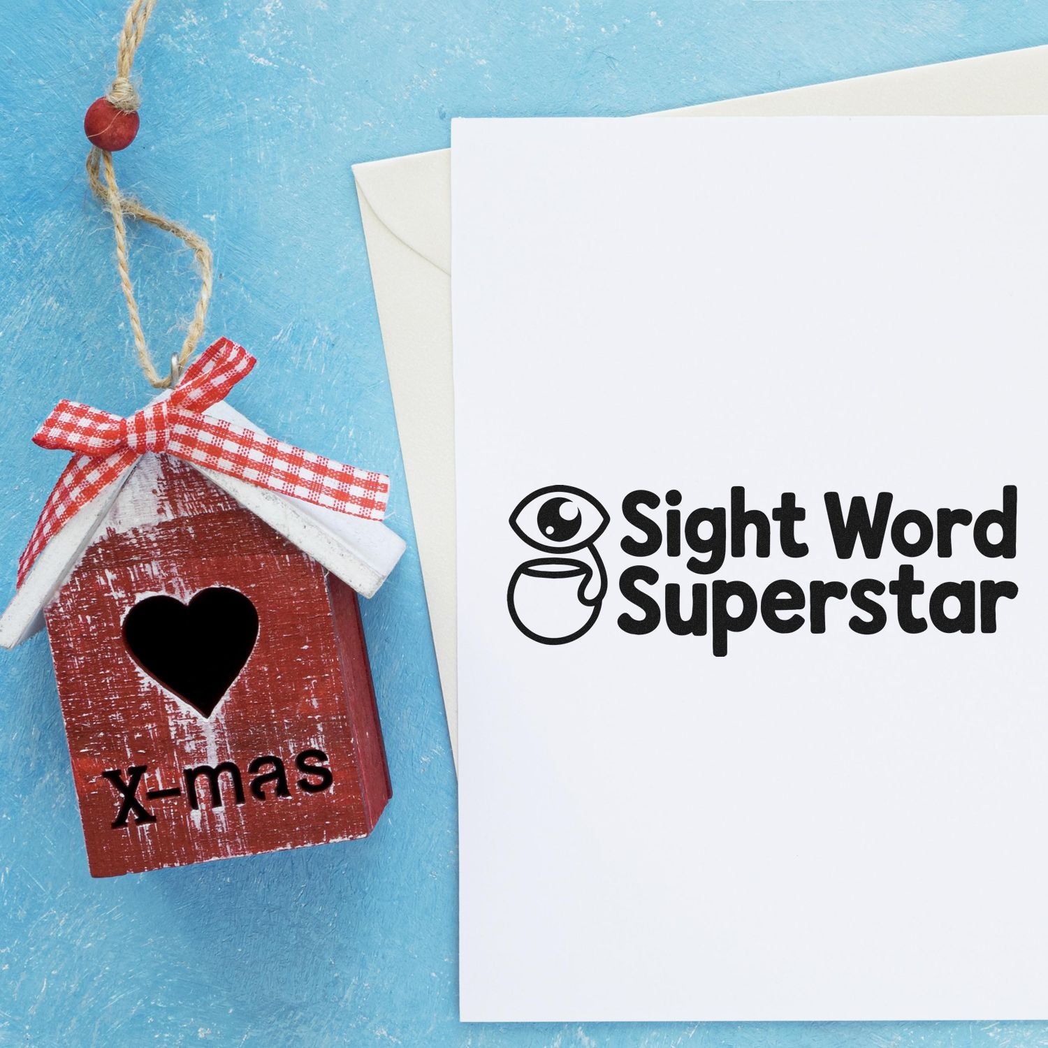 White stamp card with black Sight Word Superstar seal beside a red birdhouse ornament on a blue background.
