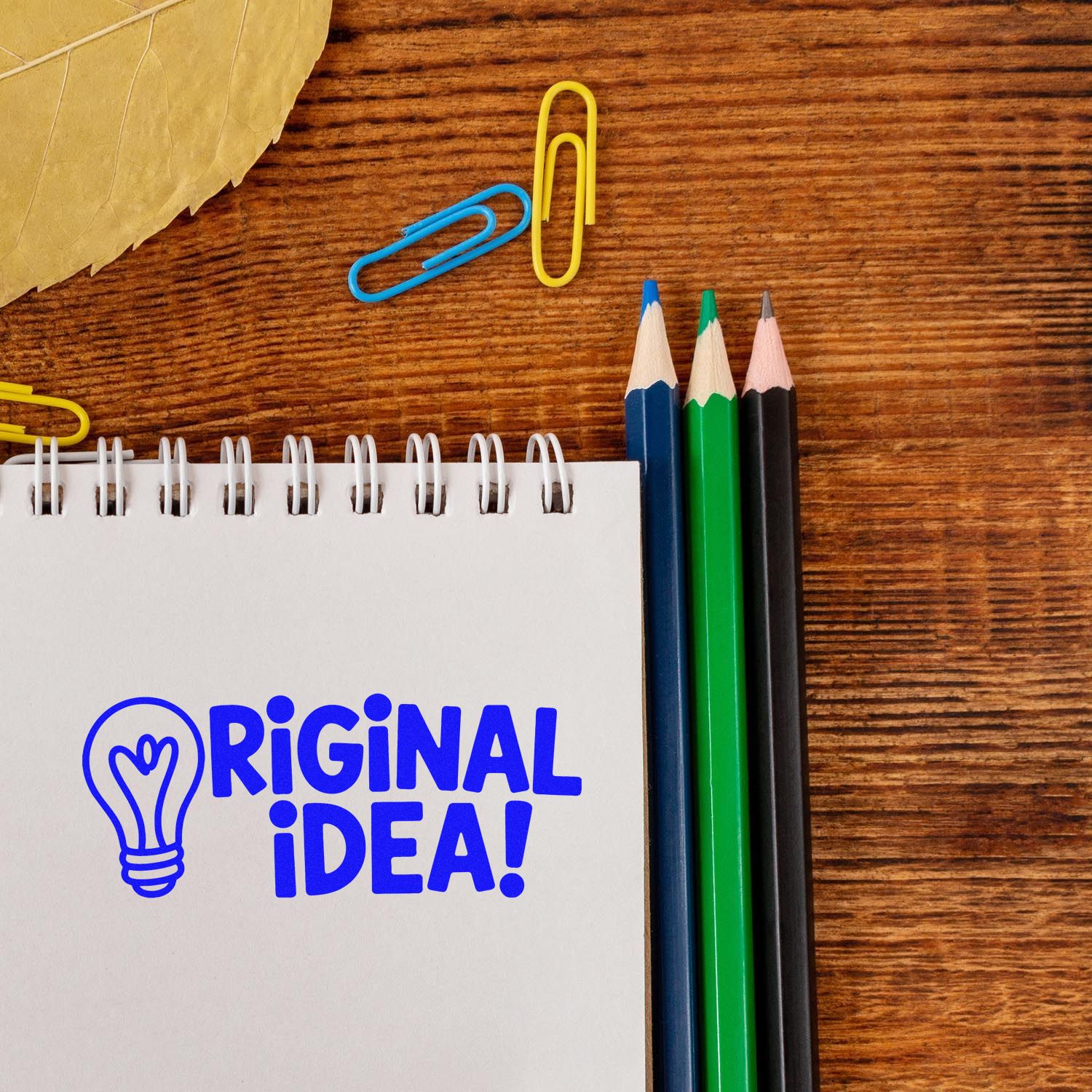 Blue ORIGINAL IDEA! stamp with light bulb on spiral notebook, beside colored pencils and paper clips on a wood desk.