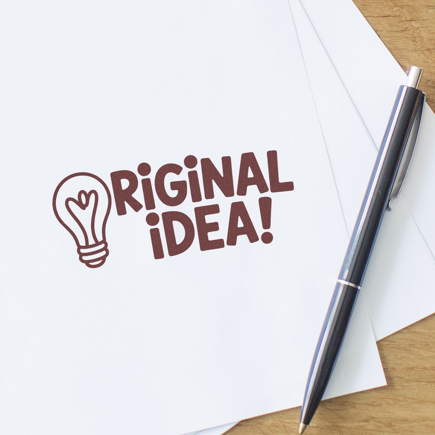 Brown ORIGINAL IDEA! stamp seal with lightbulb icon on white paper beside a black pen on a wood desk.