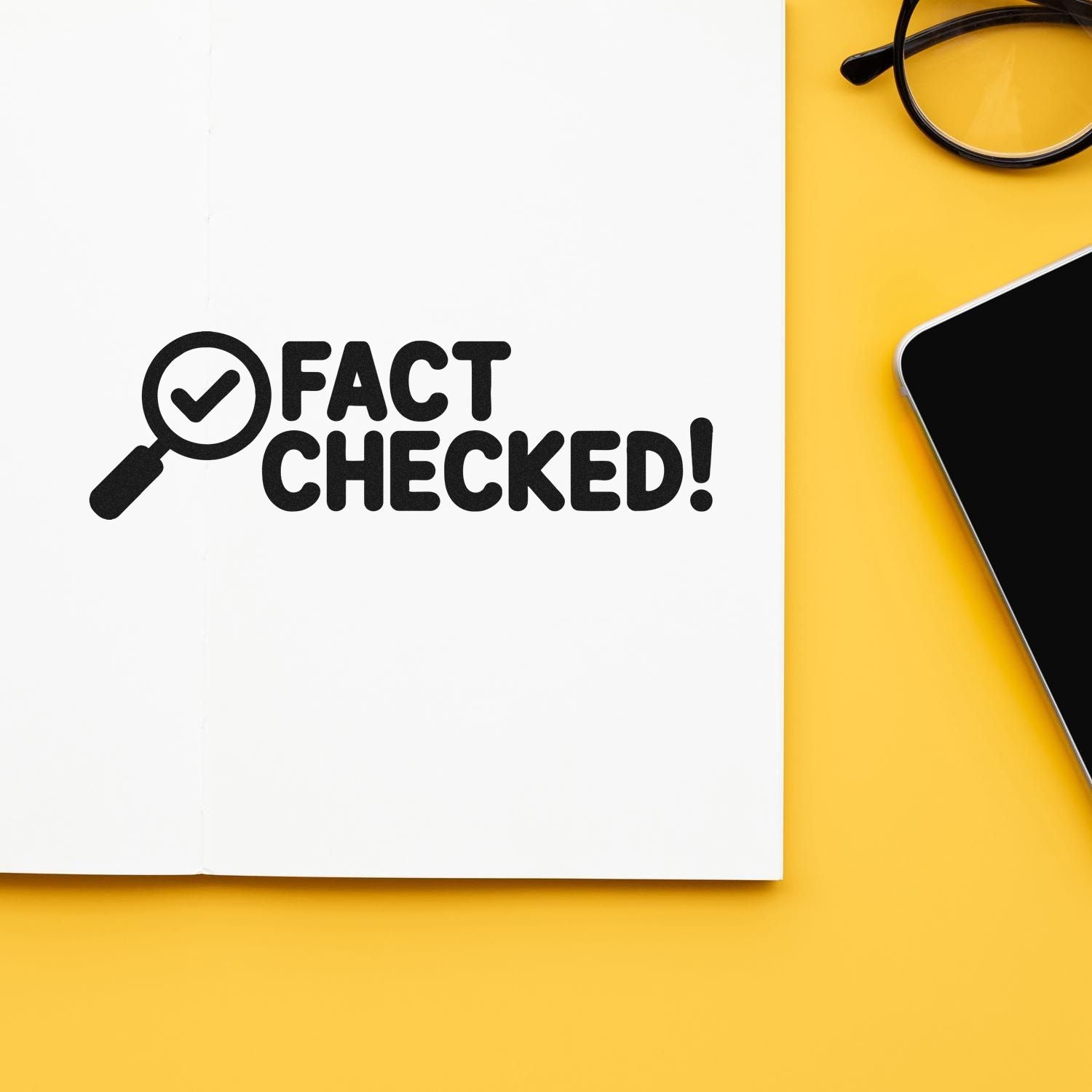 Black FACT CHECKED! stamp and seal with magnifying glass icon on white paper over a yellow desk, beside glasses and a phone.