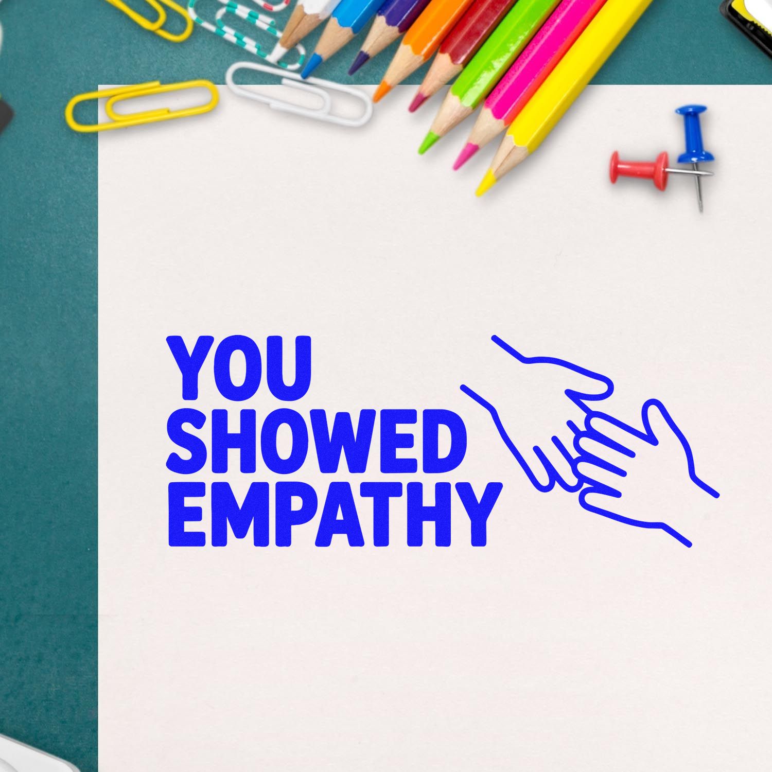 Blue empathy stamp and seal with handshake icon on white paper, surrounded by colorful pencils, paper clips, and push pins.