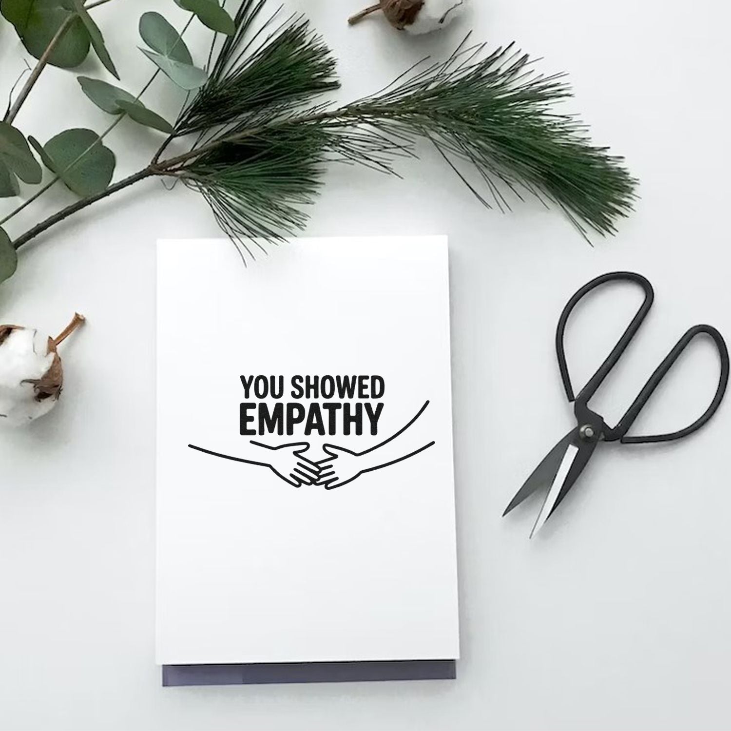 White stamp and seal card with black You Showed Empathy handshake design, beside black scissors and green foliage.