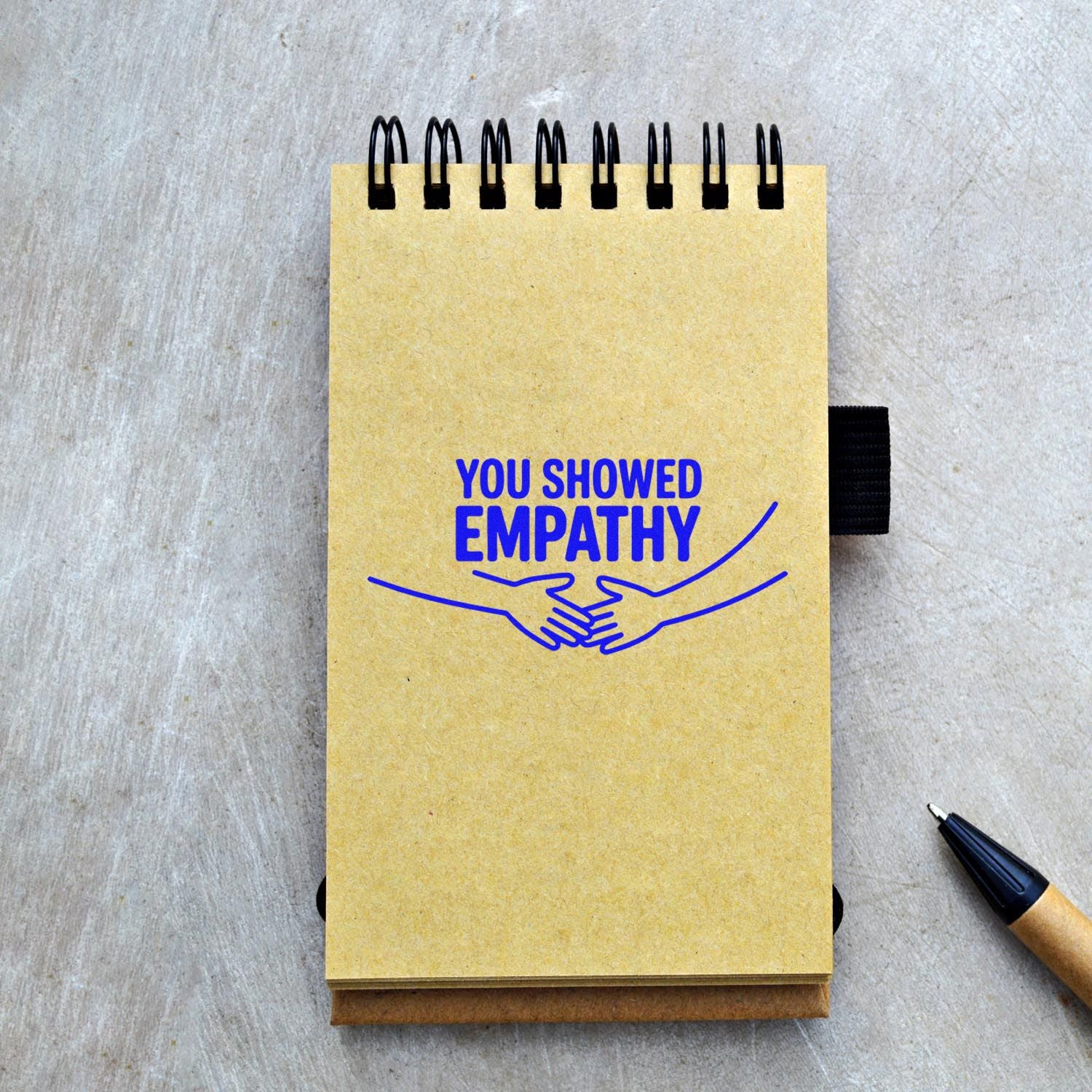 Yellow spiral notebook with blue YOU SHOWED EMPATHY stamp and handshake seal graphic, on gray wood with black pen.