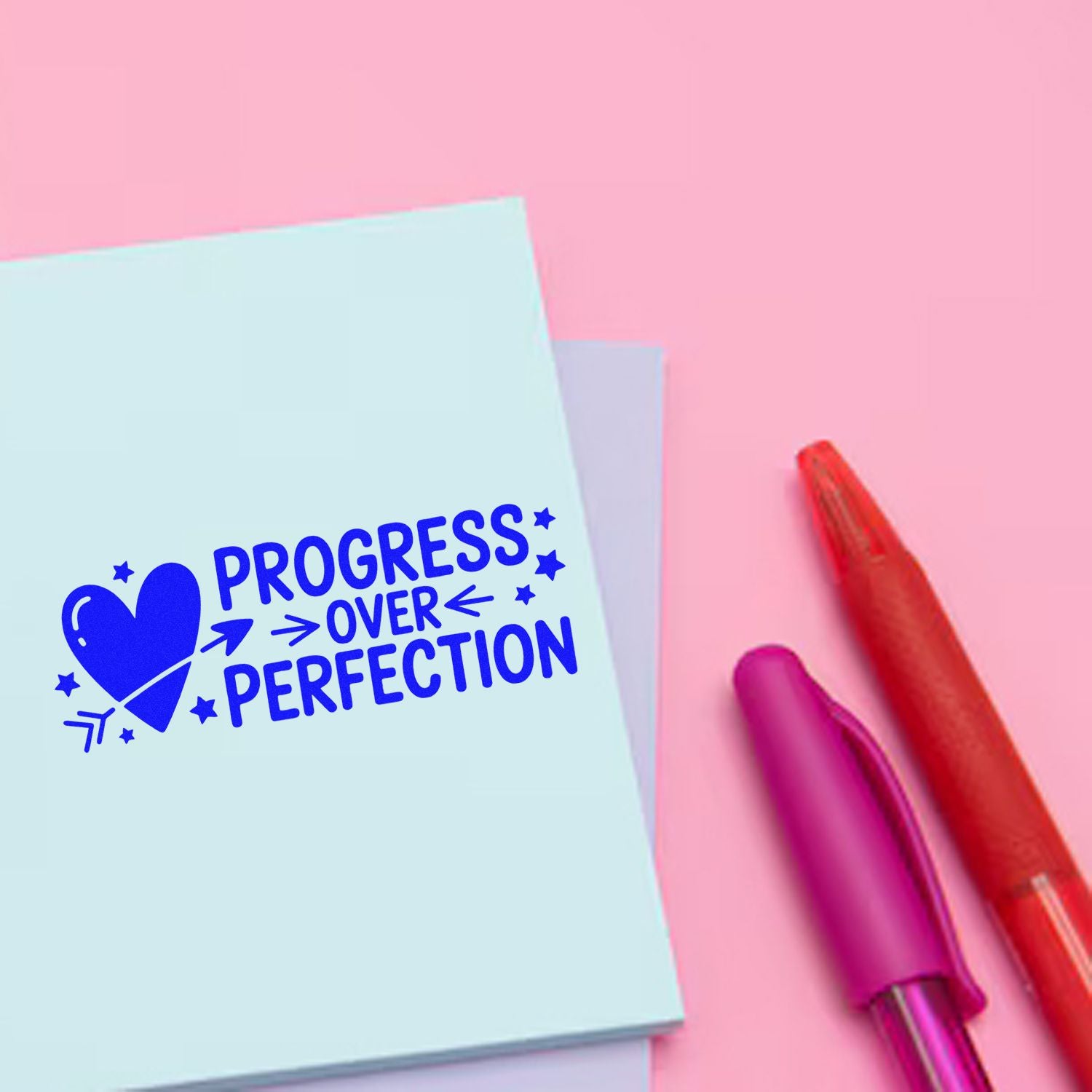 Blue progress over perfection stamp on pale blue paper beside pink and red seal pens on a pink background.