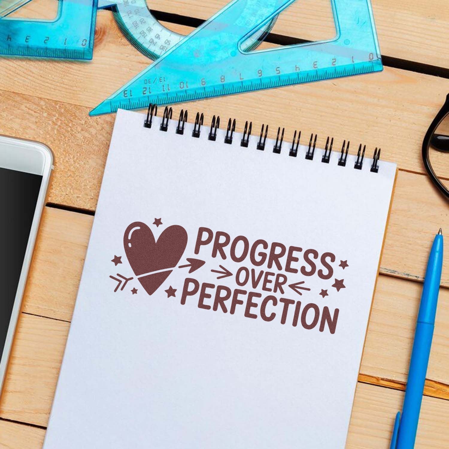 Brown progress over perfection stamp with heart on a white spiral notebook, beside blue rulers and a pen on a wooden desk.