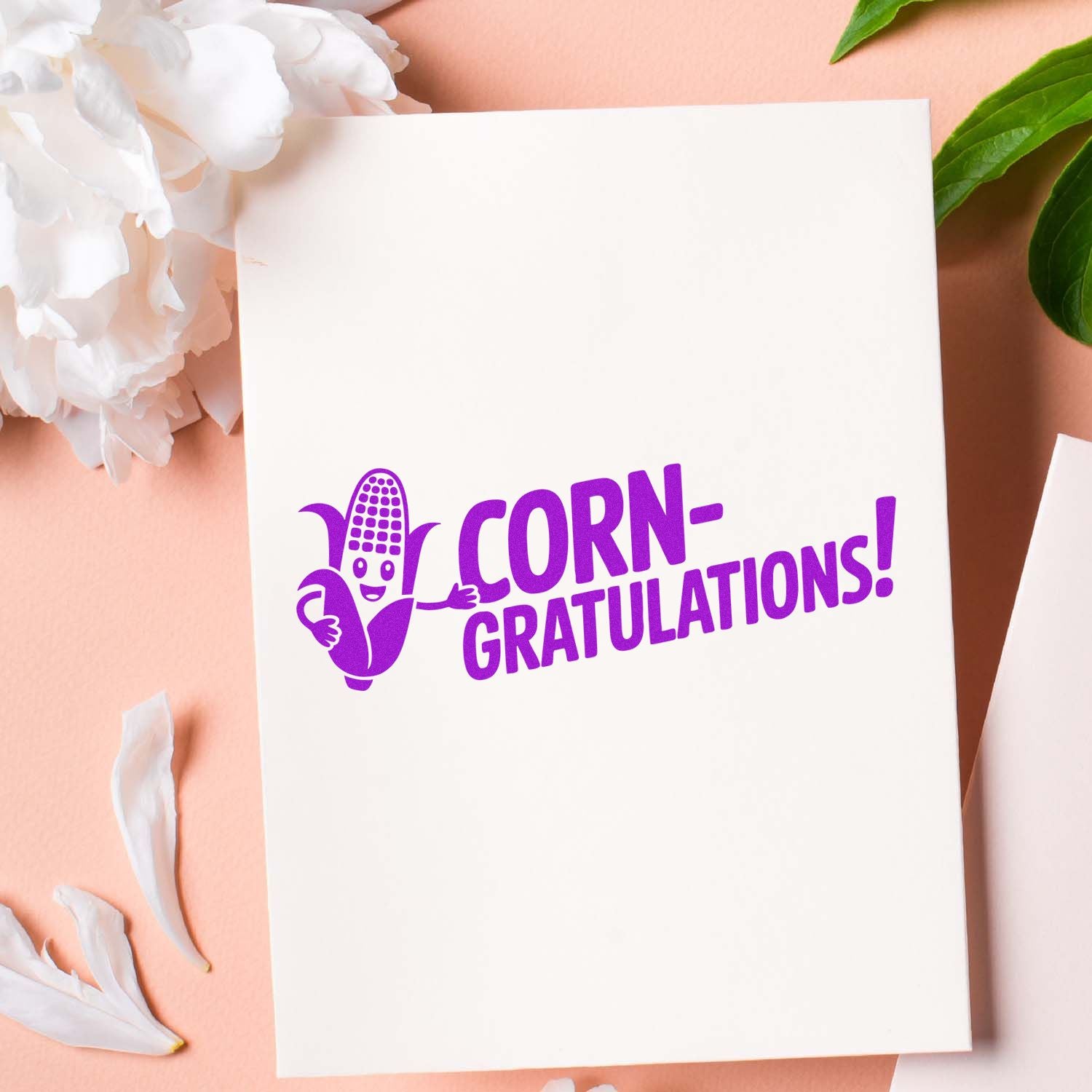 Purple Corn-gratulations stamp and seal with smiling corn graphic on a white card, on a pink background with white flowers.