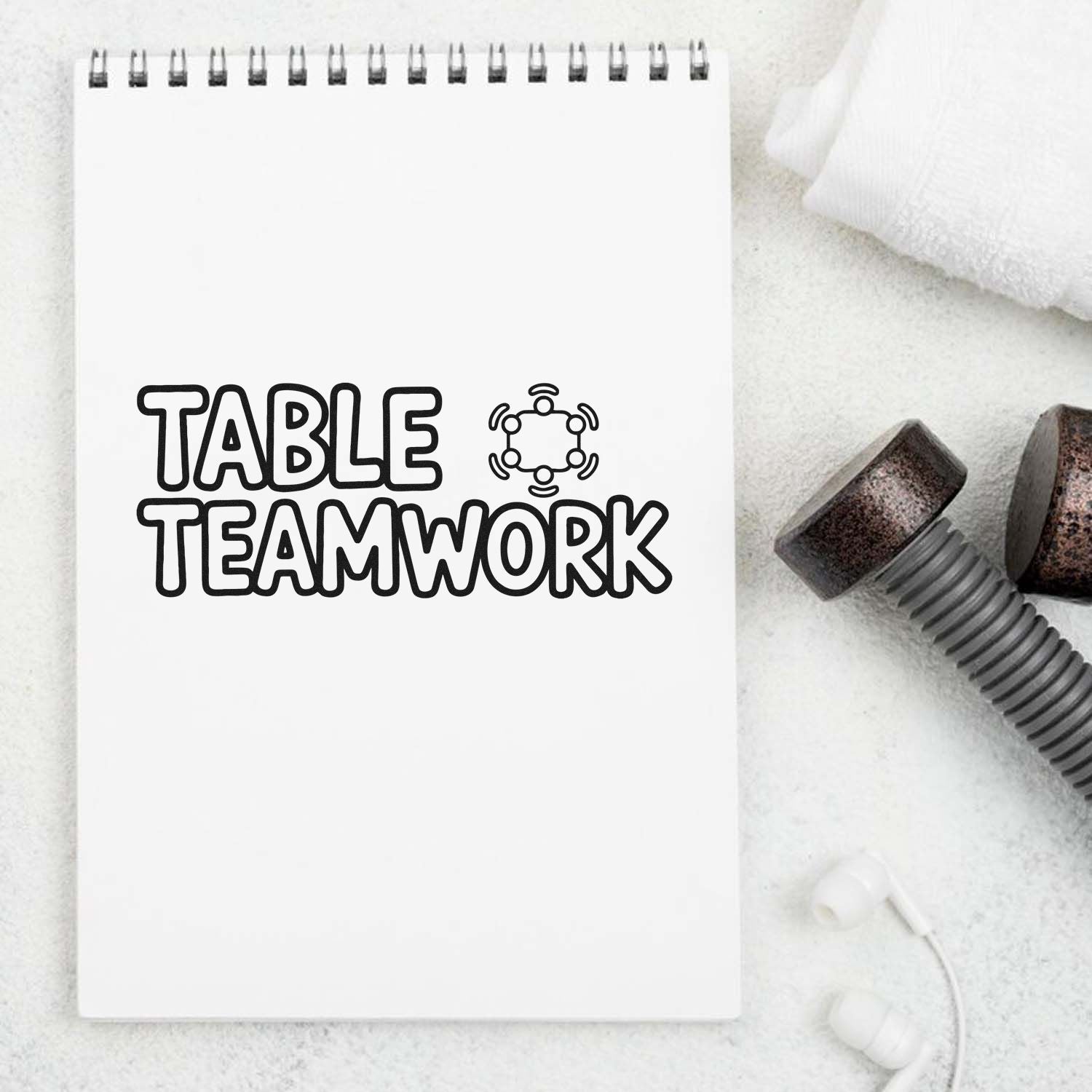 White spiral notebook with bold black TABLE TEAMWORK stamp design beside rubber seal stamps on a soft white background.