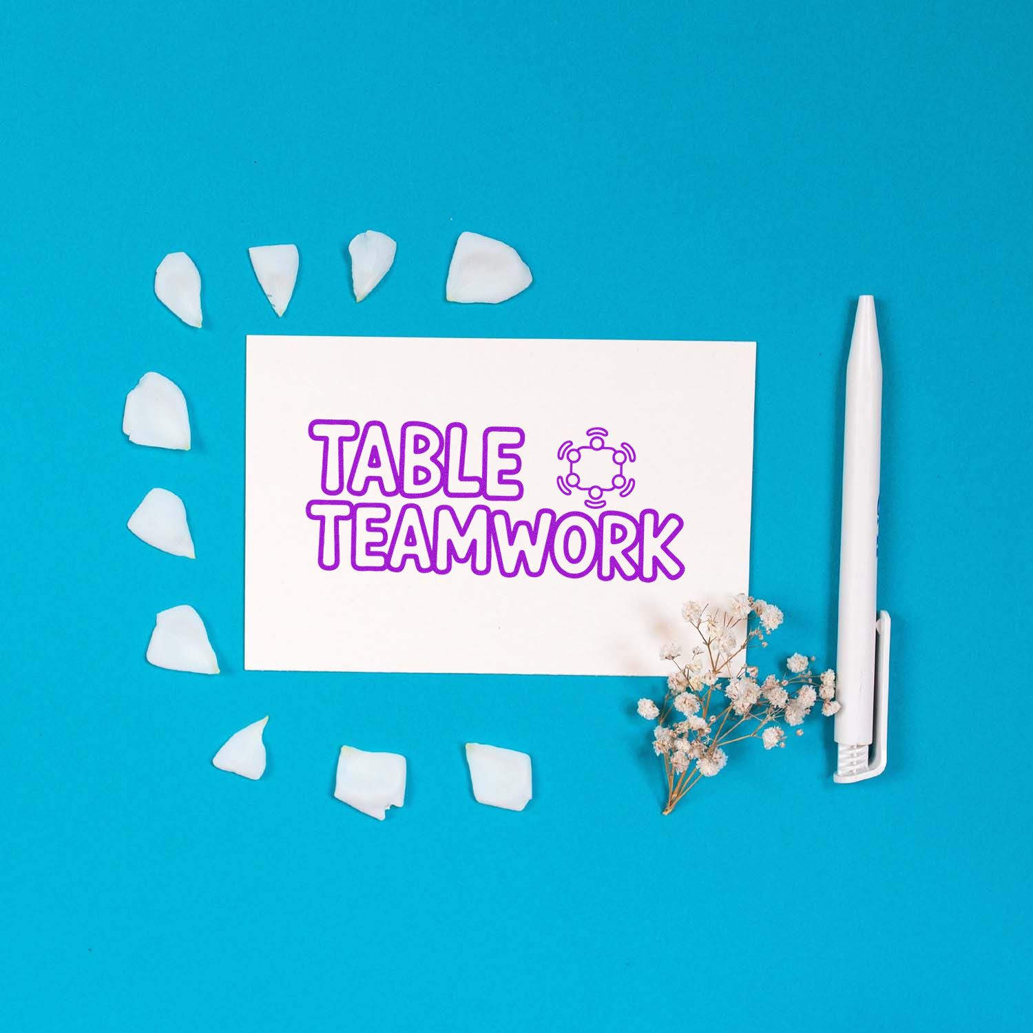 White card with purple TABLE TEAMWORK stamp, white seal petals, pen, and dried flowers on a bright blue background.