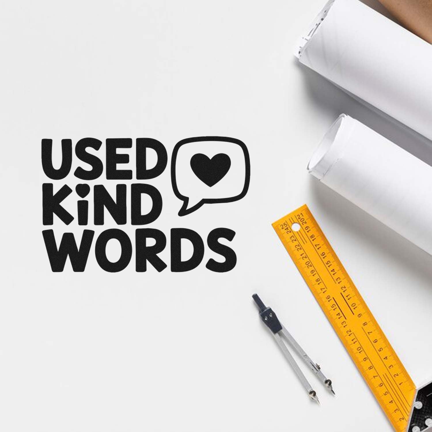 Black Used Kind Words rubber stamp with heart speech bubble, beside a yellow ruler, compass, and white paper stacks.
