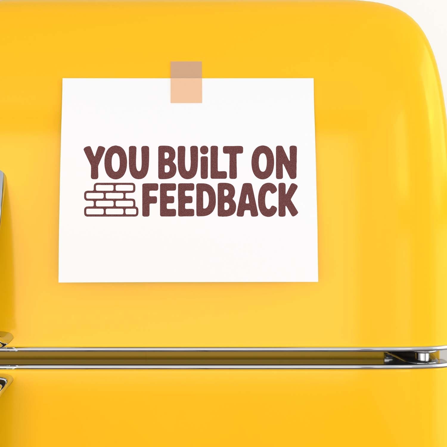 Maroon stamp-style YOU BUILT ON FEEDBACK on a white card taped to a bright yellow background, with a brick icon.