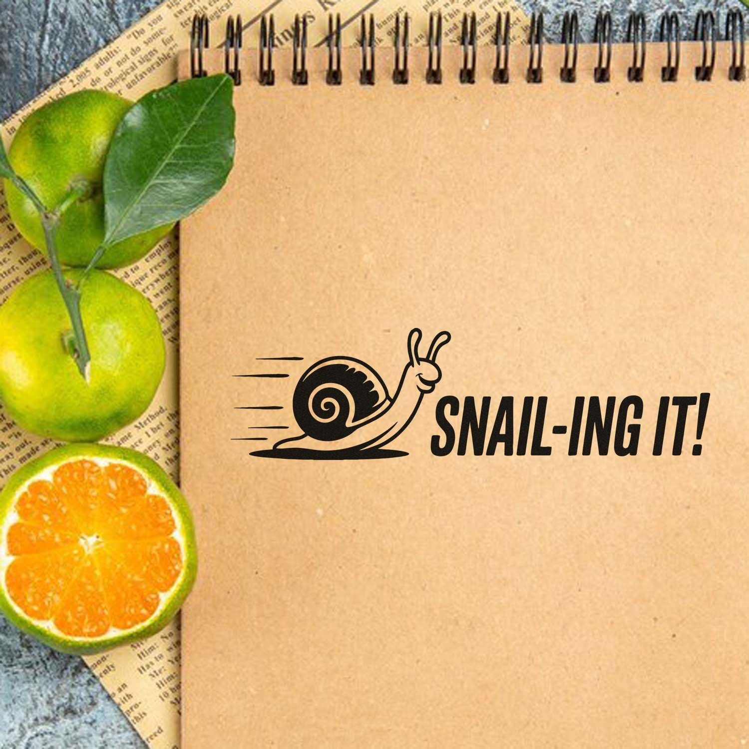 Custom stamp and seal on a kraft spiral notebook with a black snail SNAIL-ING IT! design, beside green limes and orange citrus.