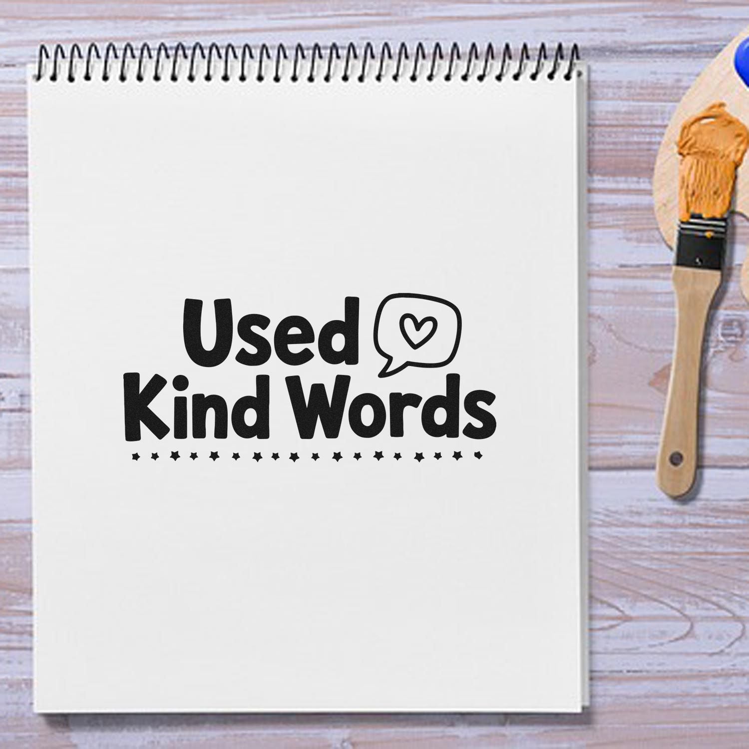Black Used Kind Words stamp on a white spiral notebook beside a wooden rubber seal stamp on a wood surface.