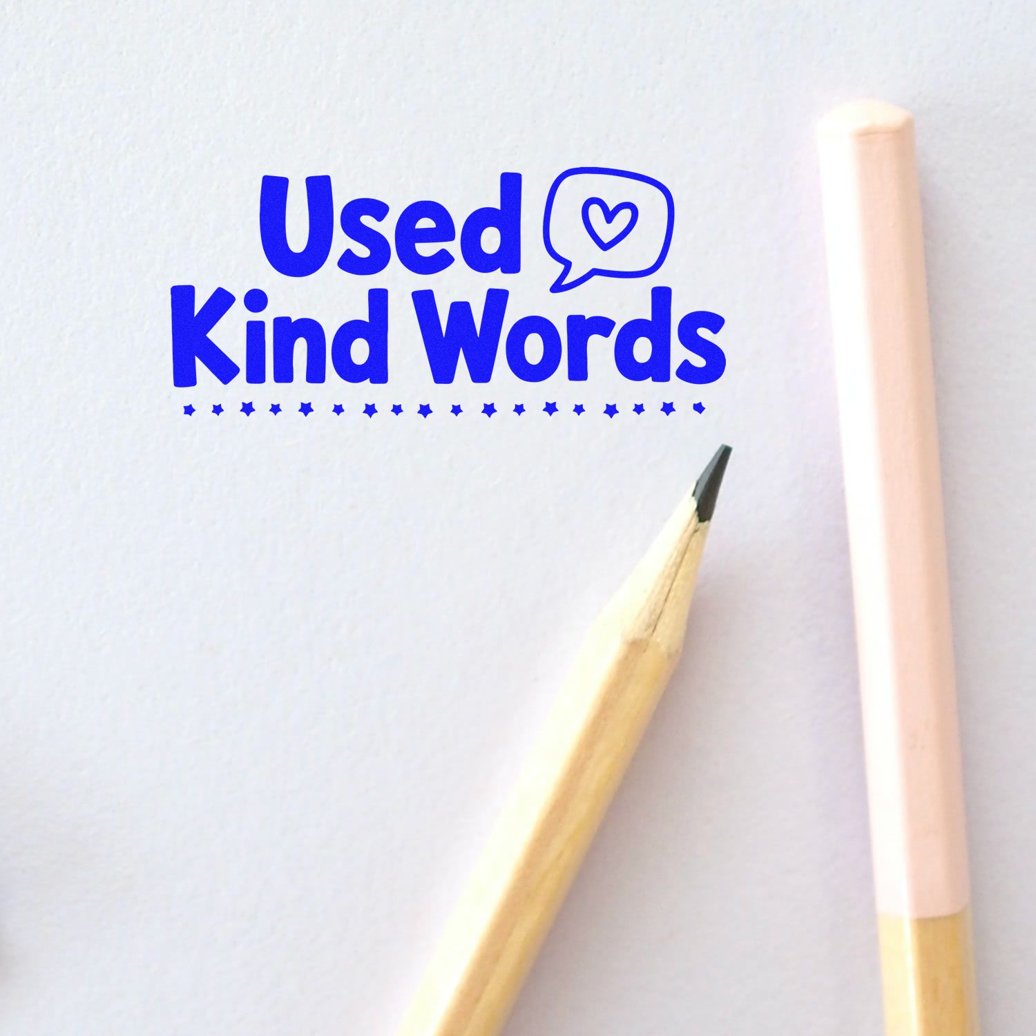 Blue Used Kind Words stamp with heart speech bubble on white paper, next to a pencil and pale pink stamp handle.