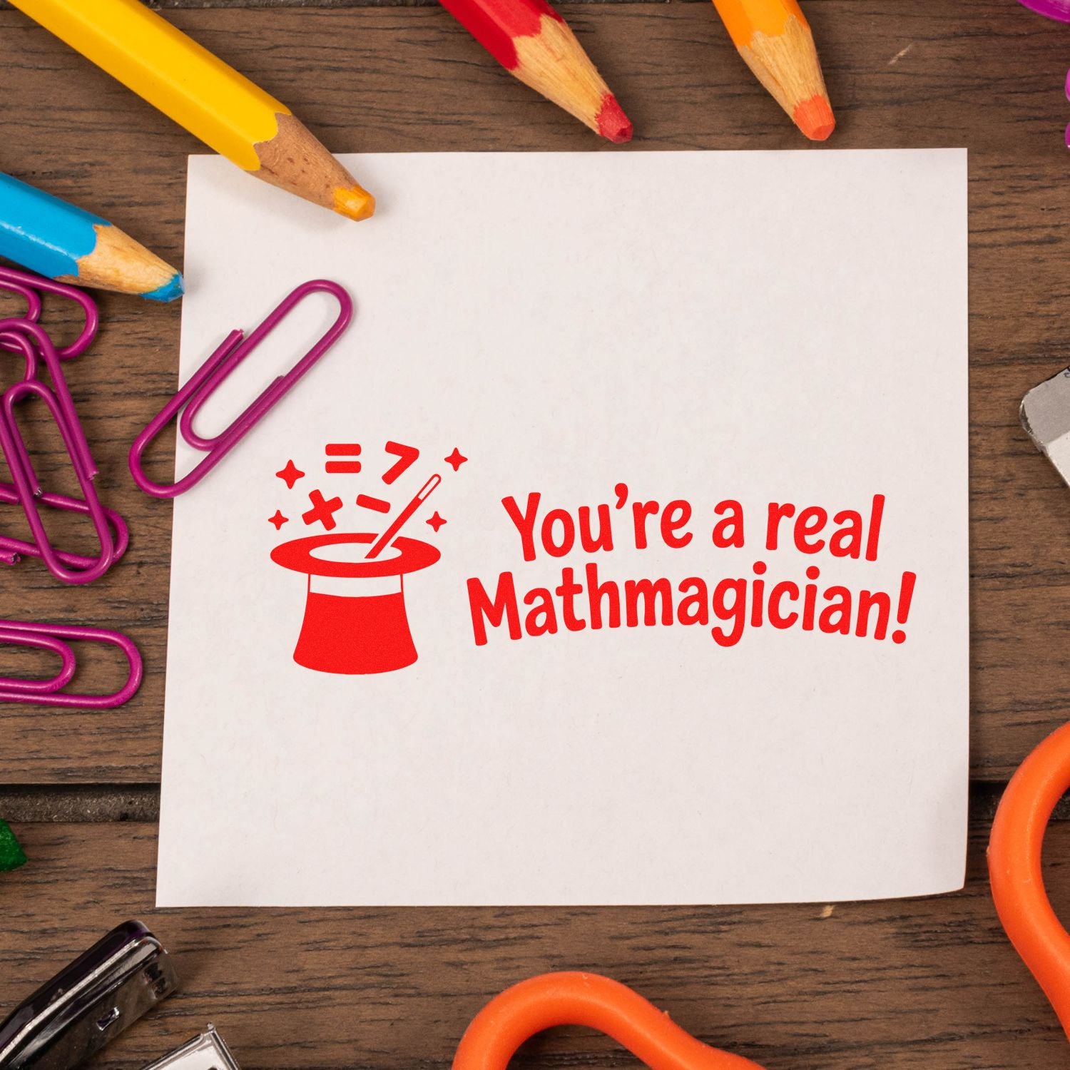 Red math magician stamp and seal on white paper, surrounded by colorful pencils and purple paper clips on wood.