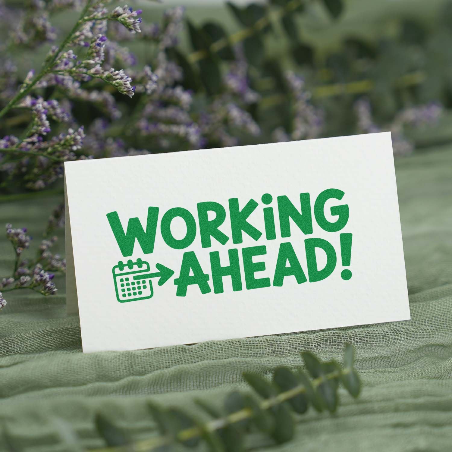 Green "Working Ahead!" stamp seal with calendar icon on white card, set among soft purple flowers and greenery.