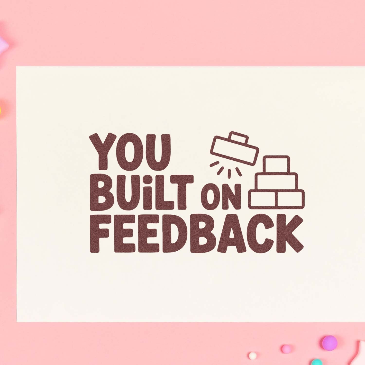 Maroon rubber stamp seal reading You Built on Feedback with stacked blocks icon on cream card over pink background.