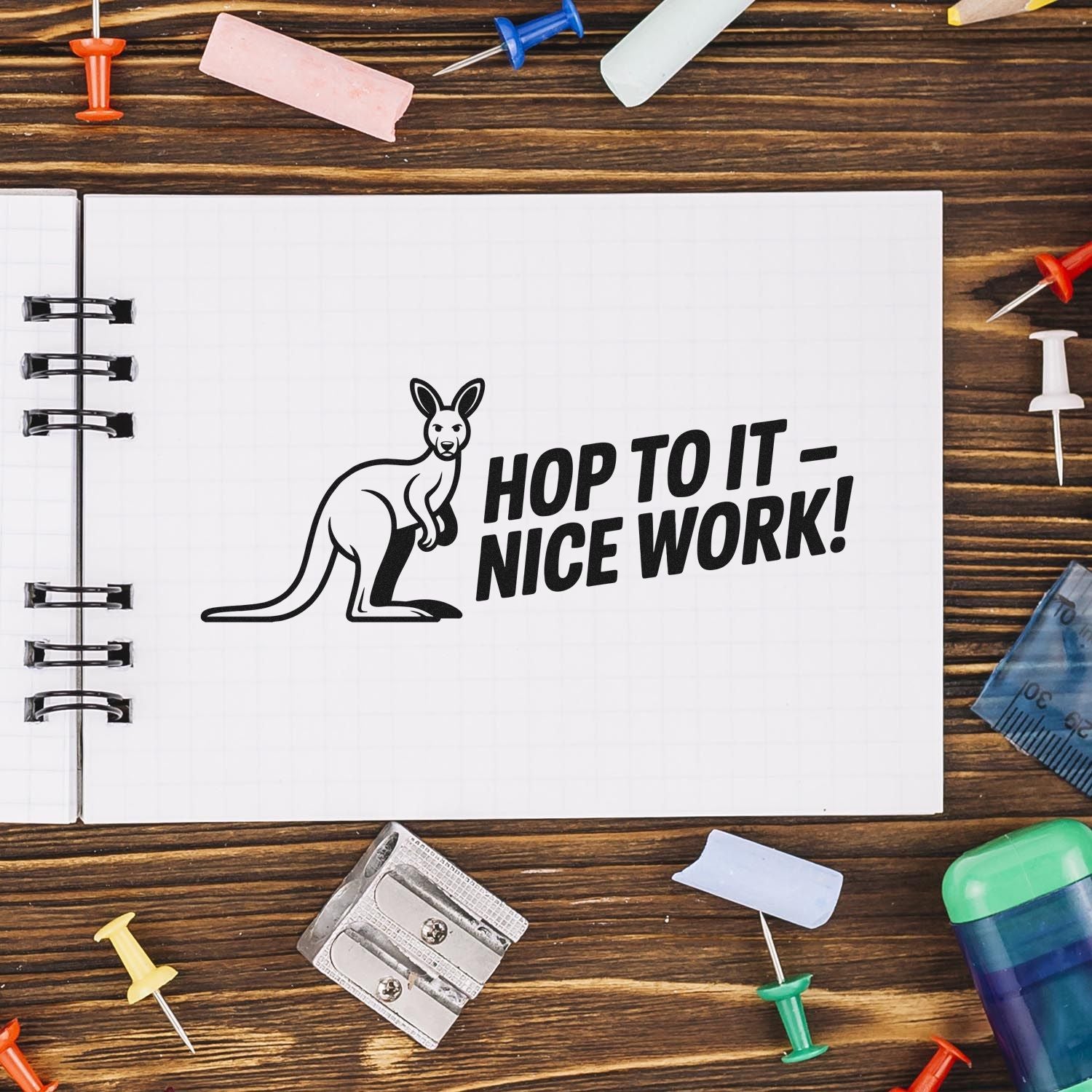 Black kangaroo rubber stamp with Hop to It  Nice Work! on a white notebook page, surrounded by colorful pushpins on wood.