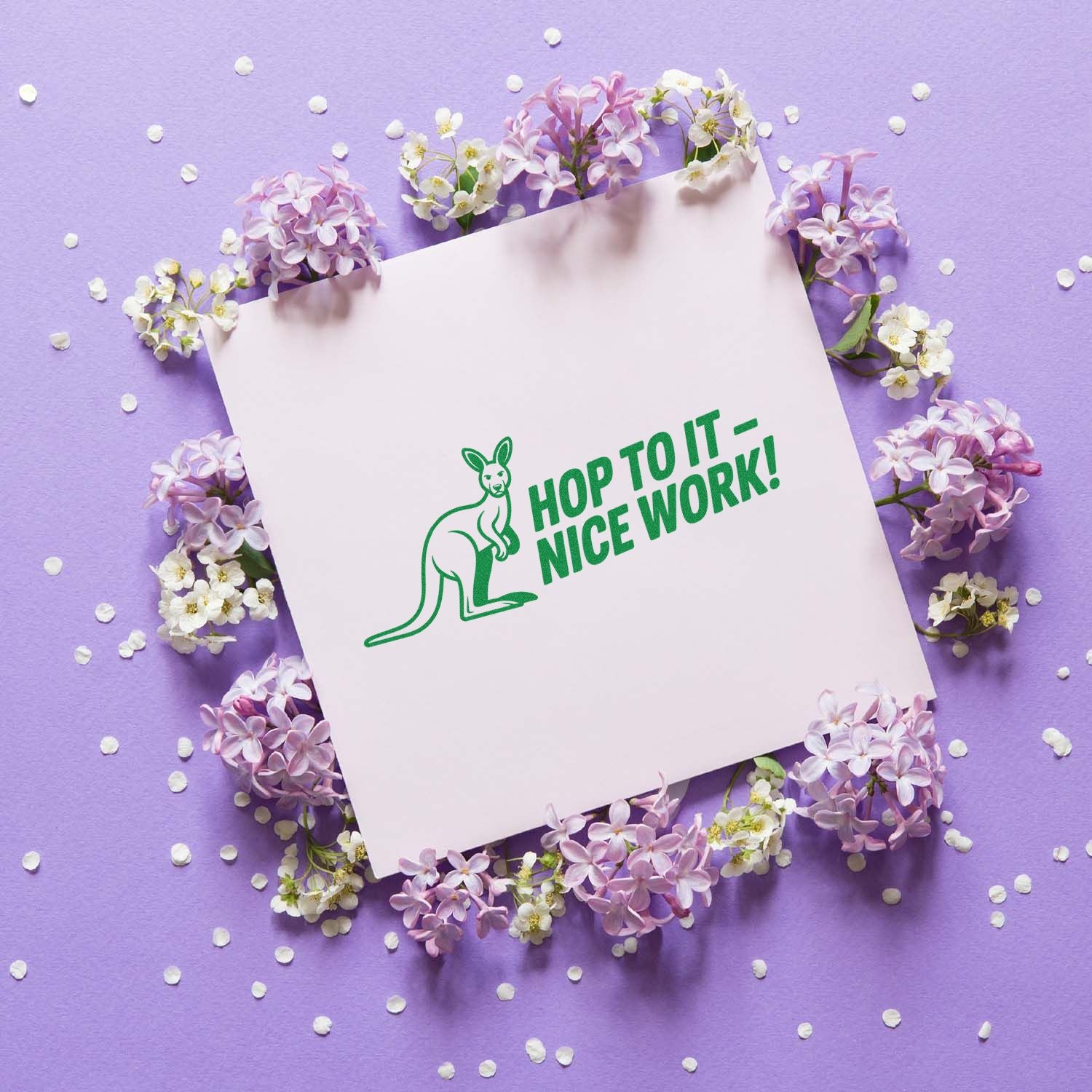 Green kangaroo rubber stamp seal reading Hop to it - Nice work! on white card, with lilac flowers on lavender background.