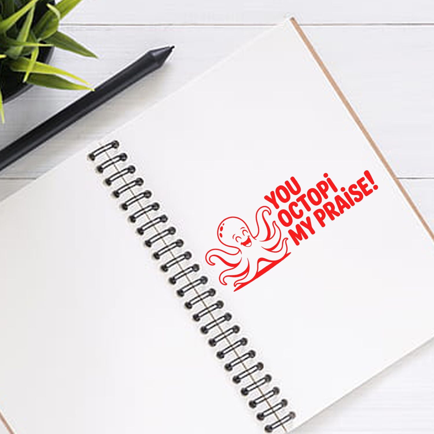 Red octopus rubber stamp seal on a white spiral notebook, with black pen and green plant.