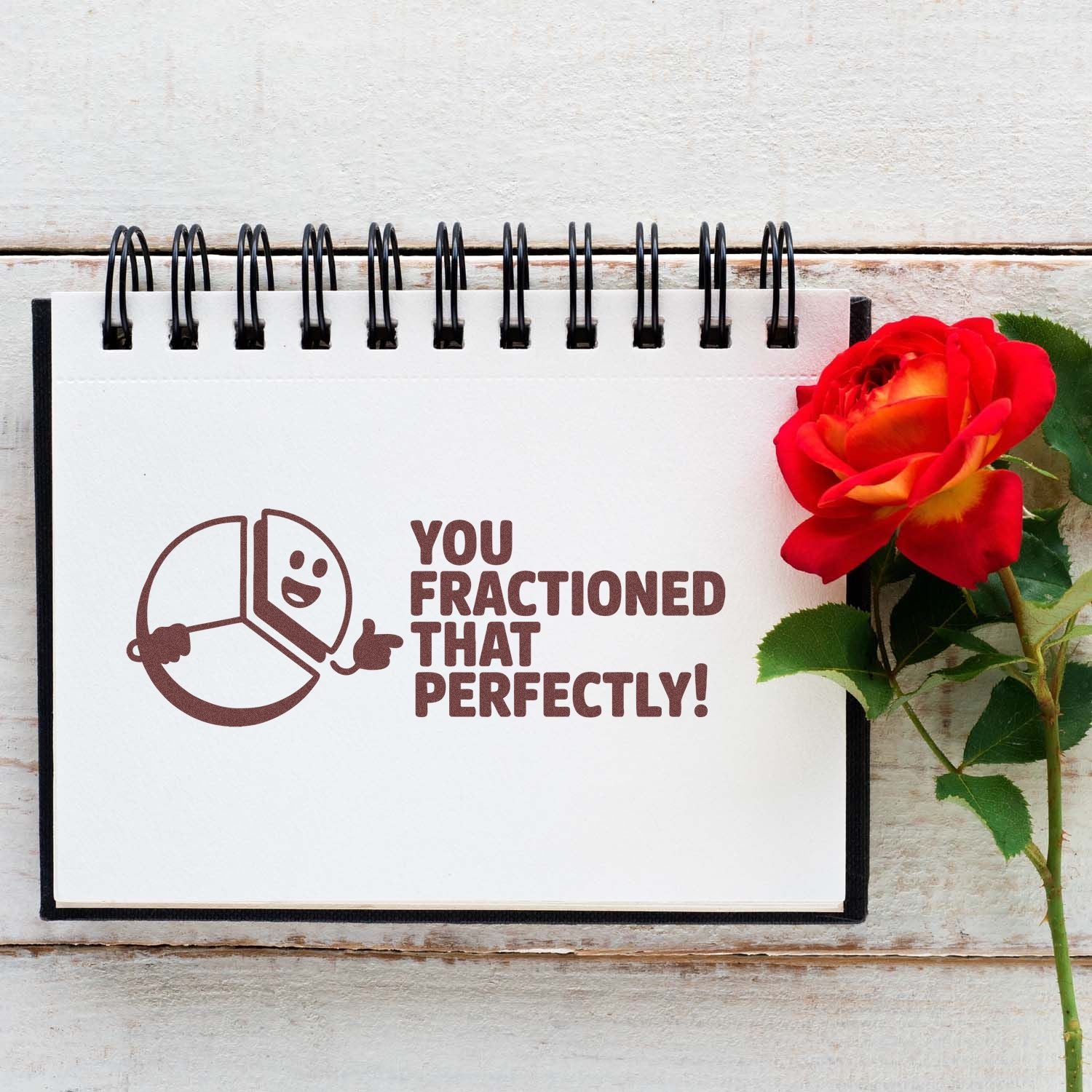 Brown rubber stamp/seal design on a white spiral notebook reading 'You fractioned that perfectly!' beside a red rose with green leaves.