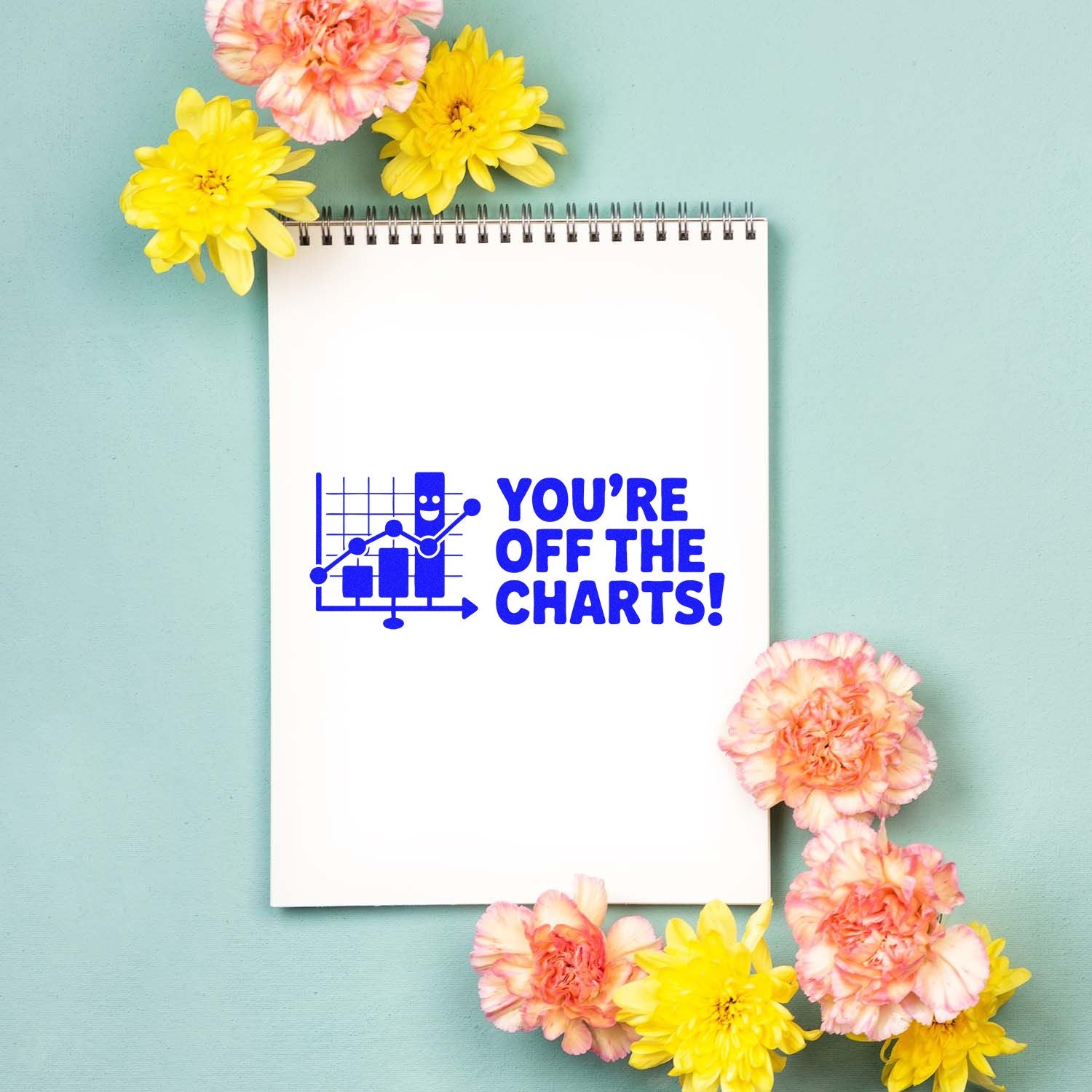 White spiral notebook with blue stamp seal reading Youre off the charts! on mint background, framed by pink and yellow flowers